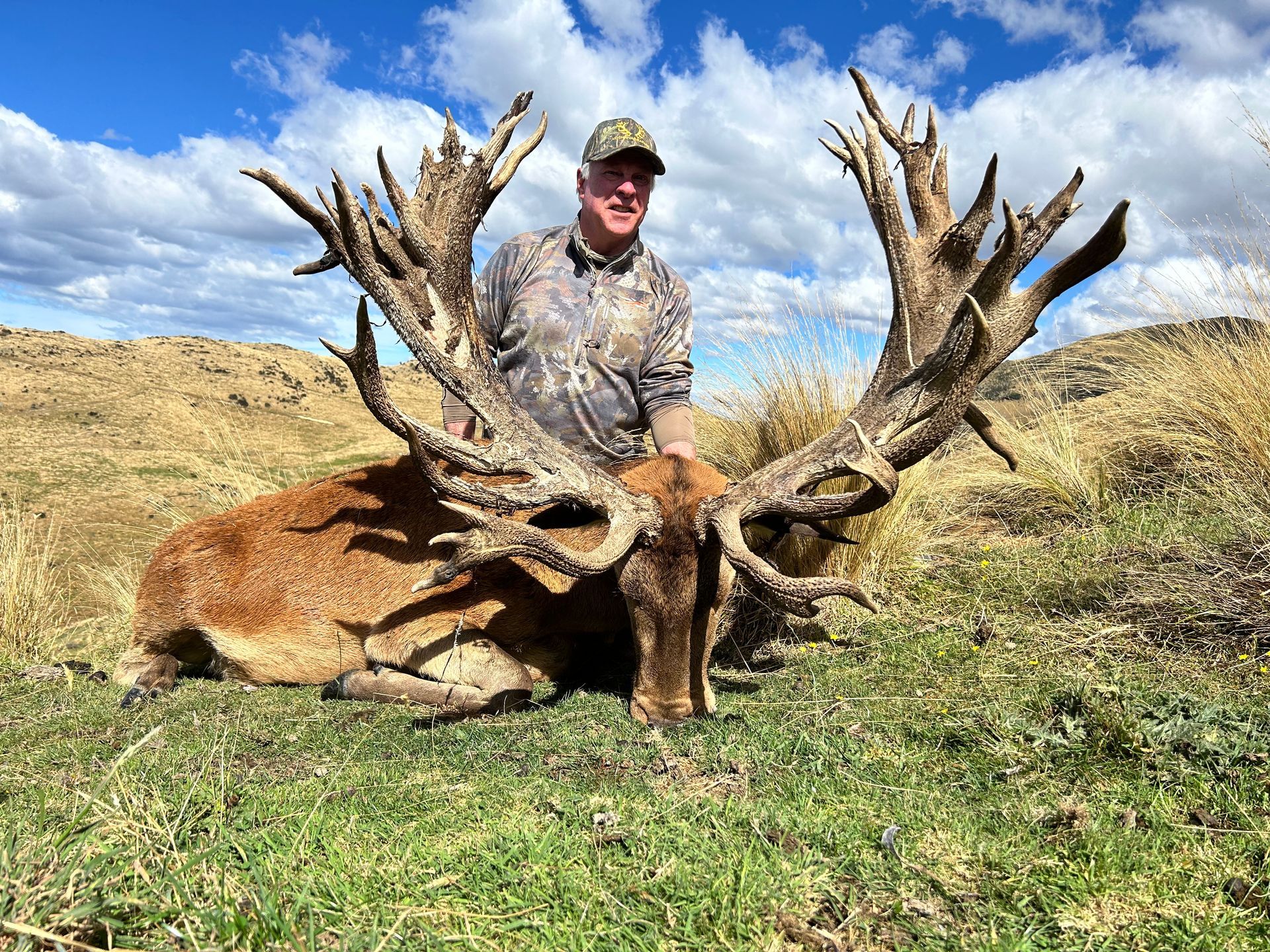 A man in camouflage poses with a large red deer with massive antlers on a grassy hillside under a blue sky.