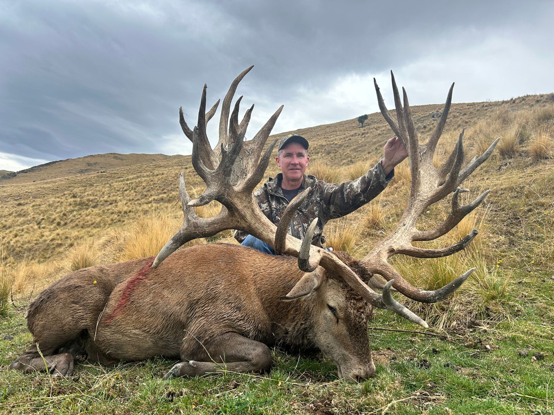 Man in camo sits beside a large dead stag with enormous antlers on a grassy hillside under an overcast sky.