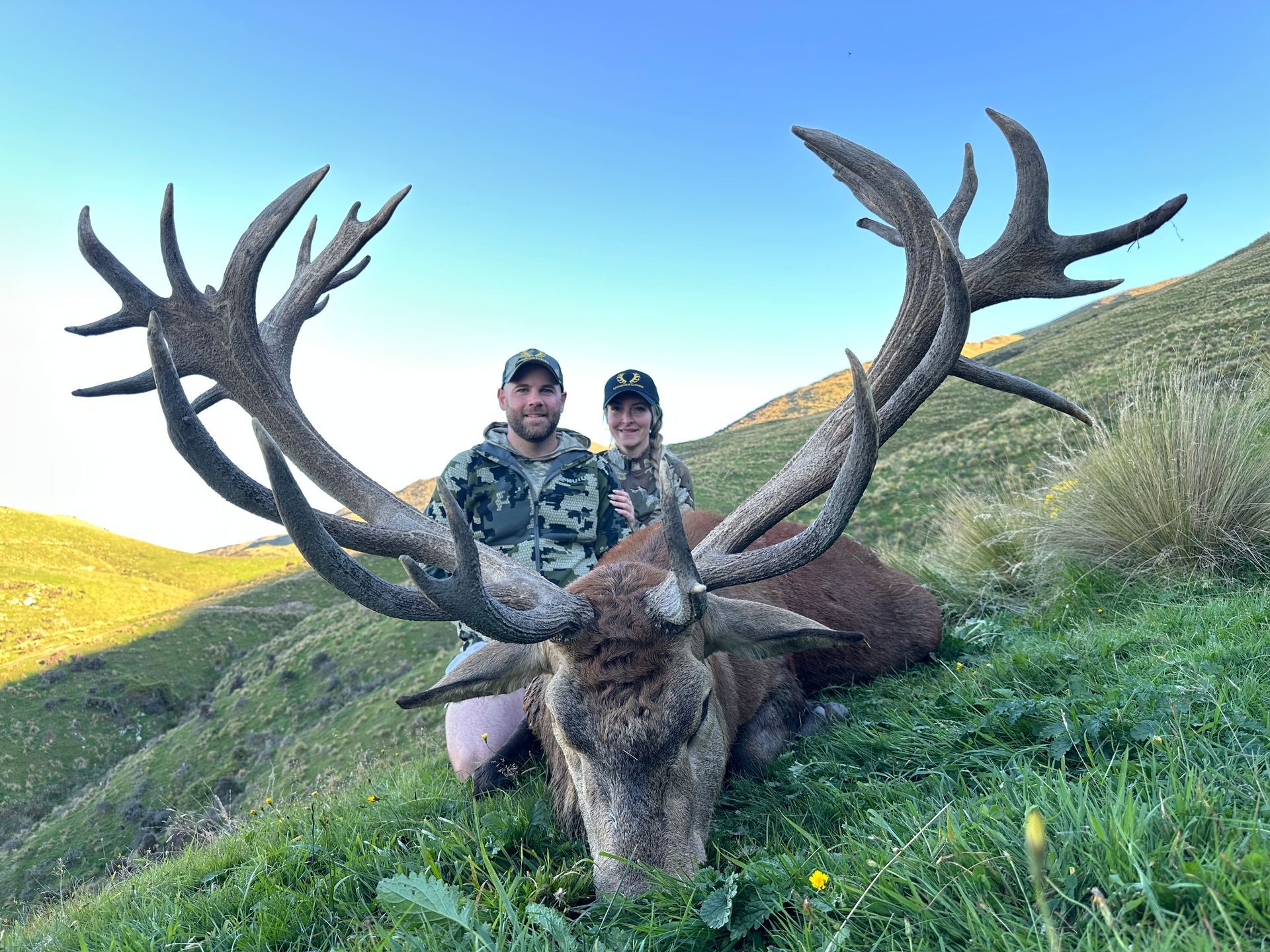 Two people pose with a large red deer stag lying in grass on a hillside under a clear blue sky. The man wears camouflage.