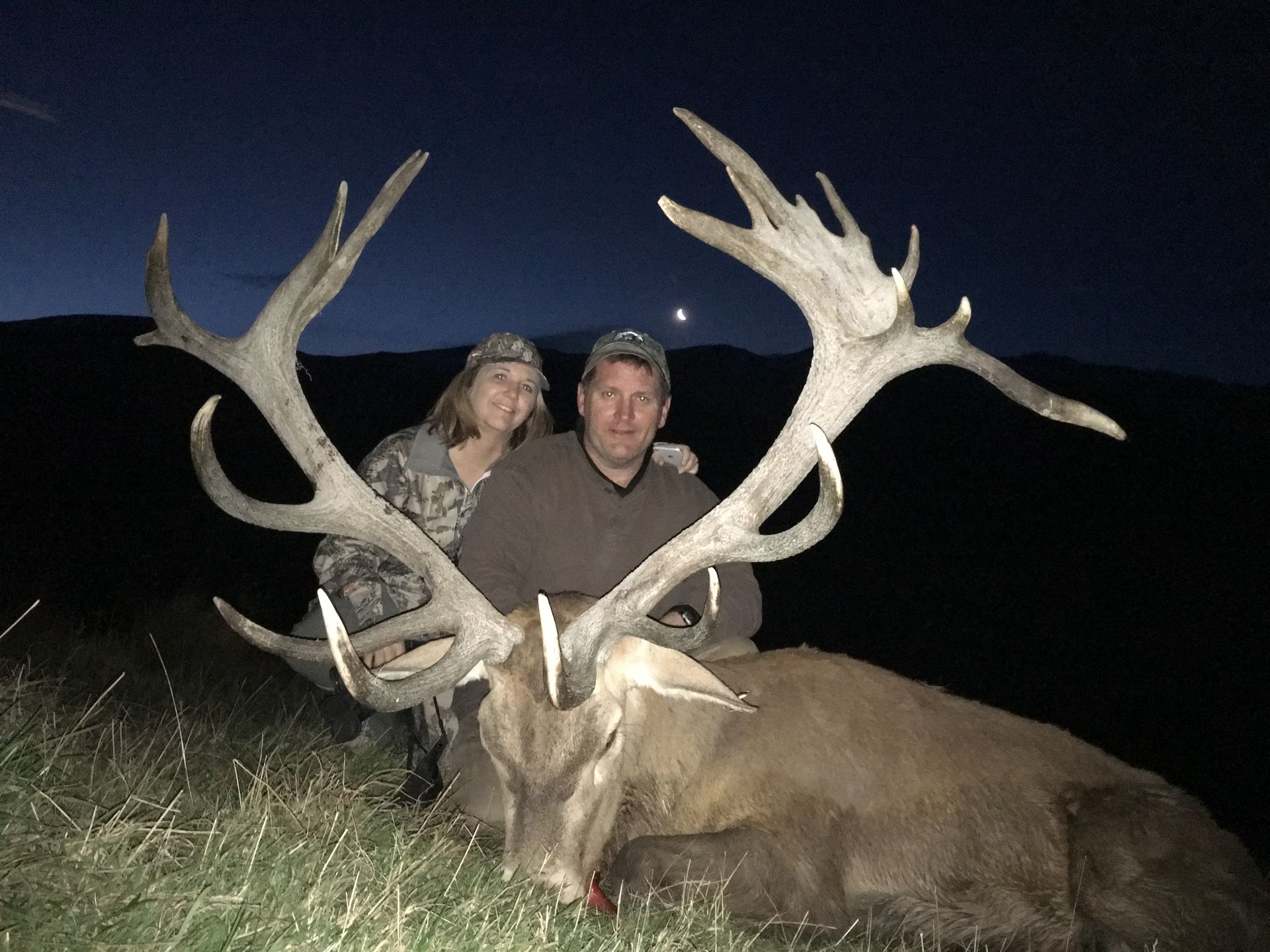 A man and woman pose with a large deer, antlers dominating the frame, set against a dark night sky. They appear to be celebrating a successful hunt.