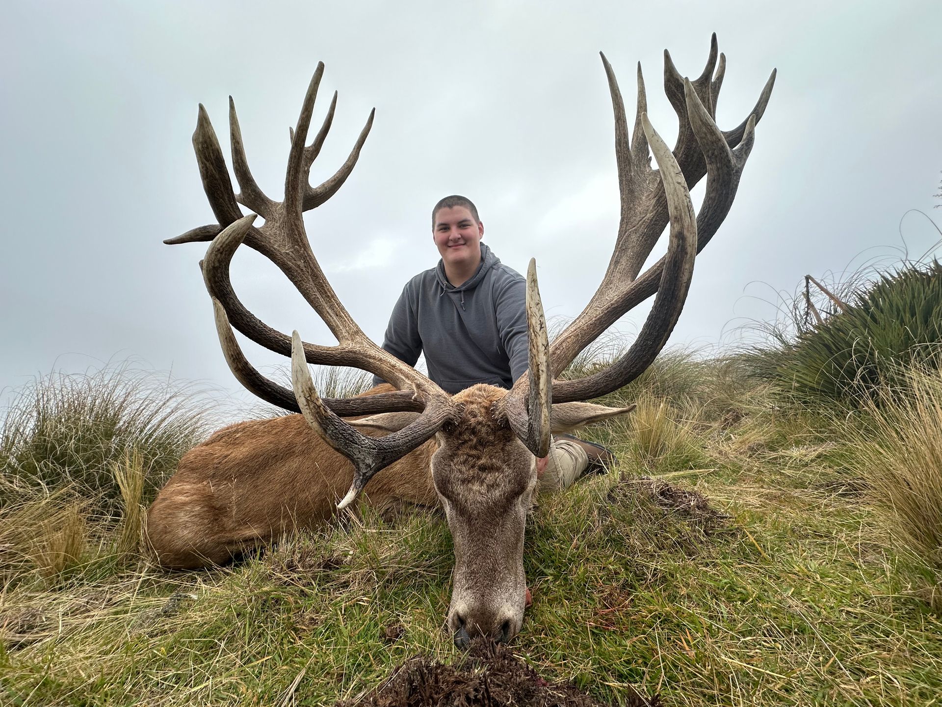 Man posing with a large dead red deer, antlers spread wide. He's smiling on a grassy hillside, overcast sky.