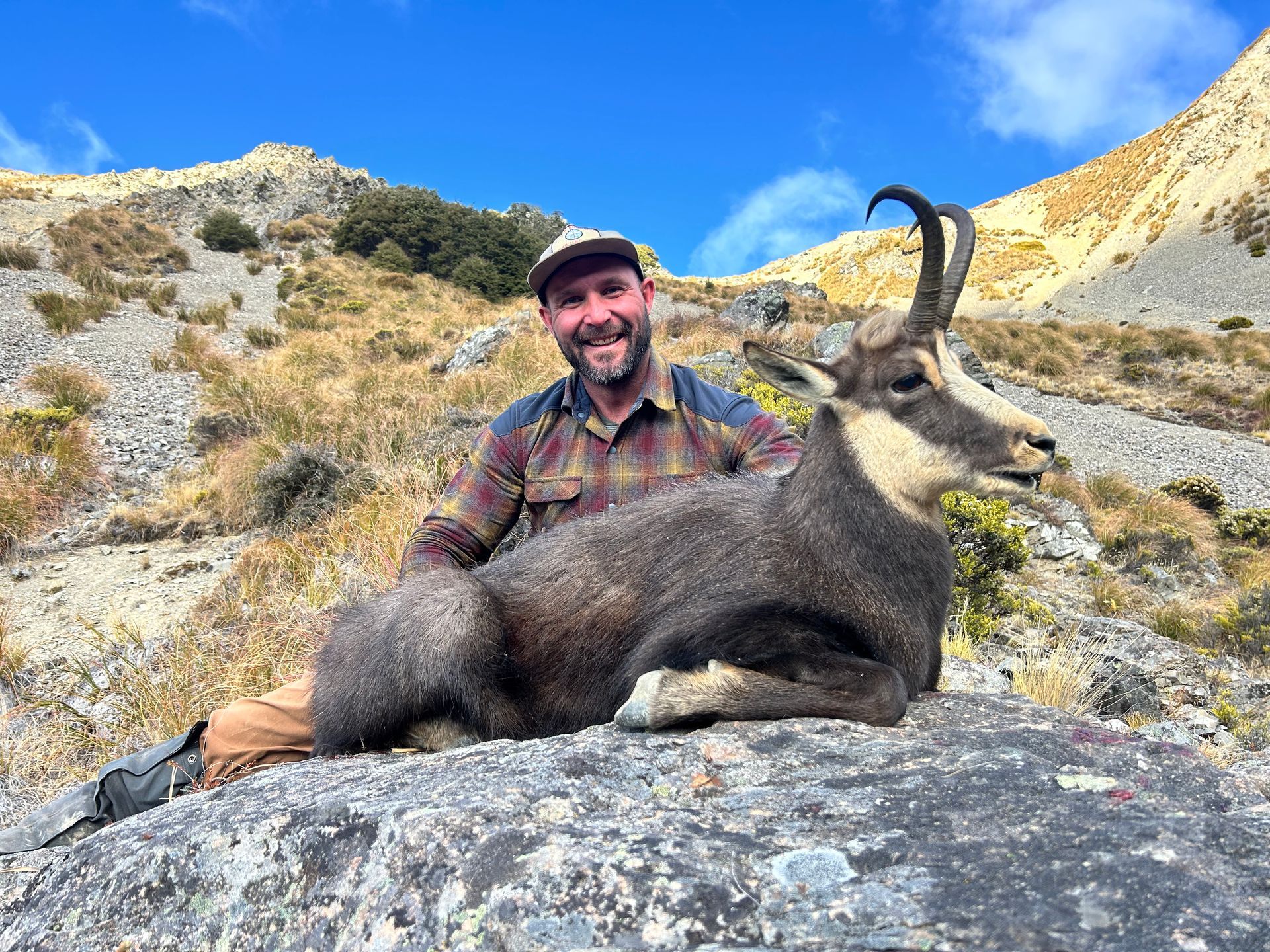 Man smiles, kneeling next to a large, dark-furred chamois with curved horns in a rocky, mountainous landscape.