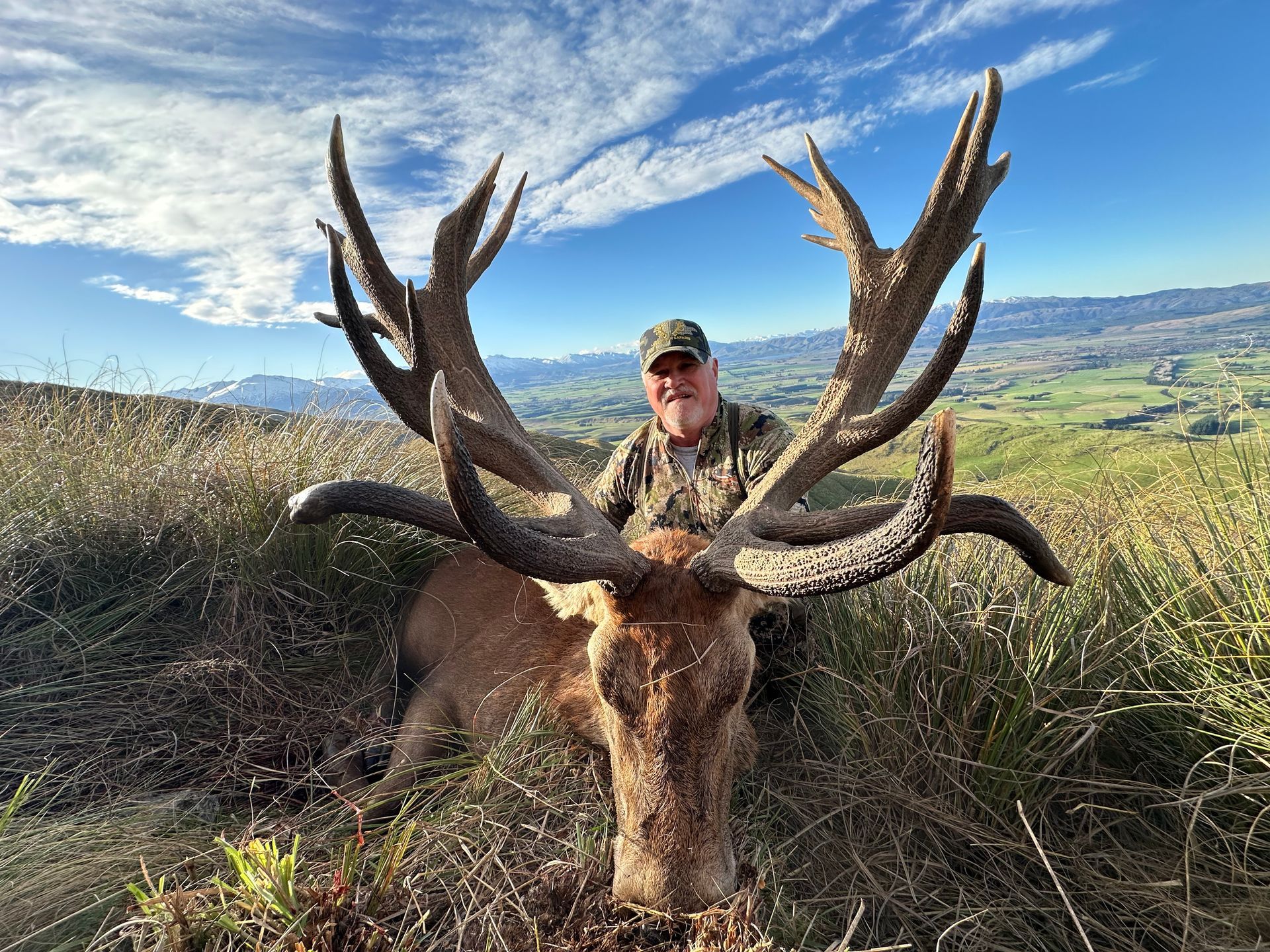 Man in camouflage stands behind a large stag with impressive antlers in a grassy field under a blue sky.
