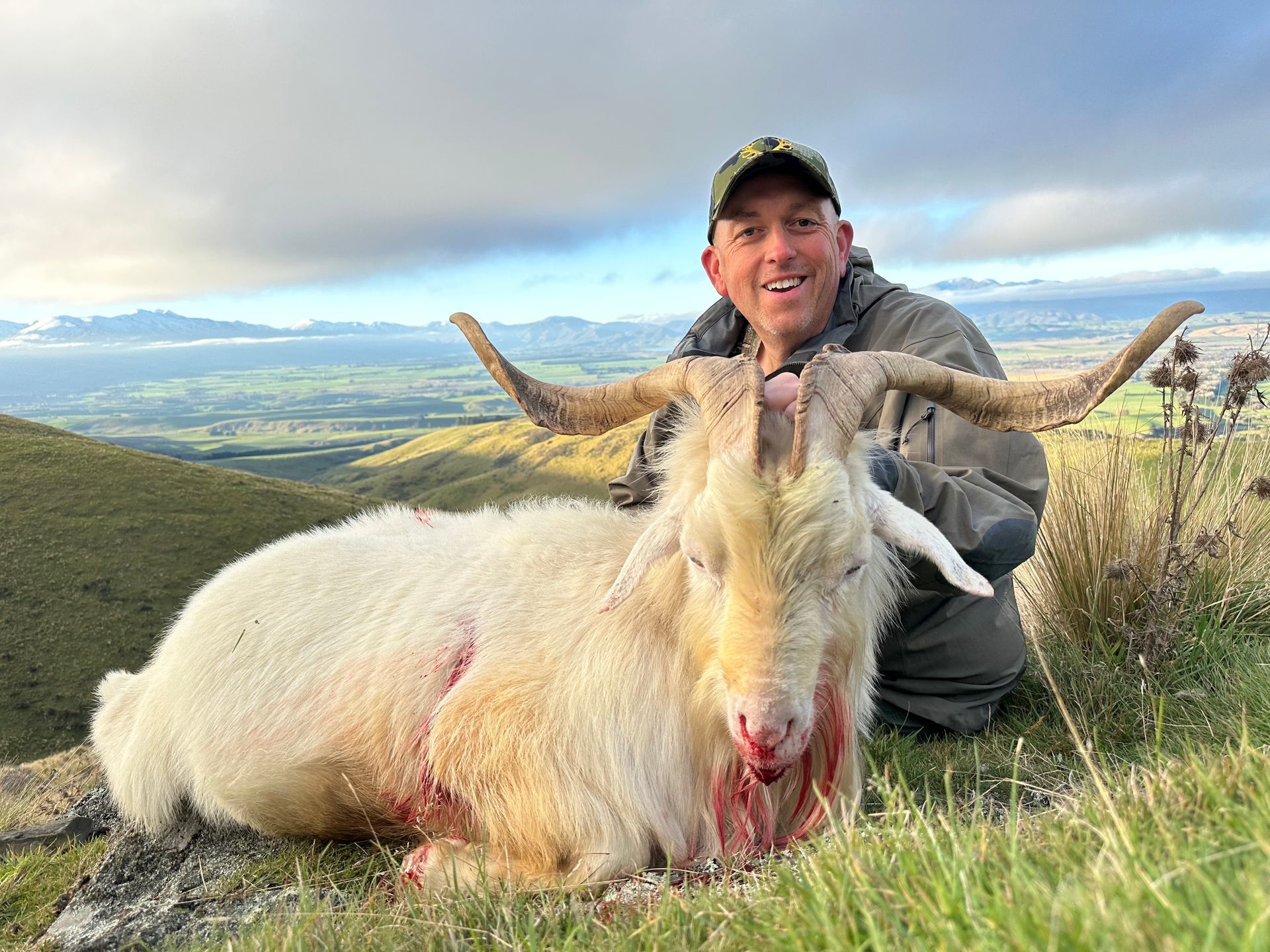 Man kneels beside a large, white goat with impressive horns, smiling in a grassy, mountain landscape. The goat shows signs of a recent kill.