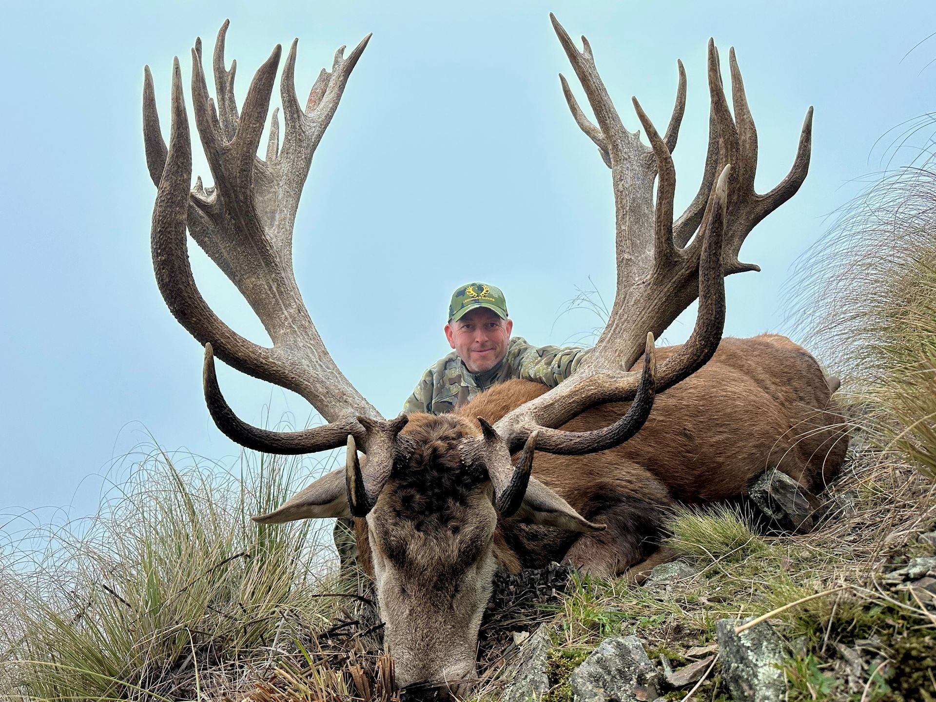 A man in camouflage poses behind a large, dead red deer with massive antlers; outdoors.