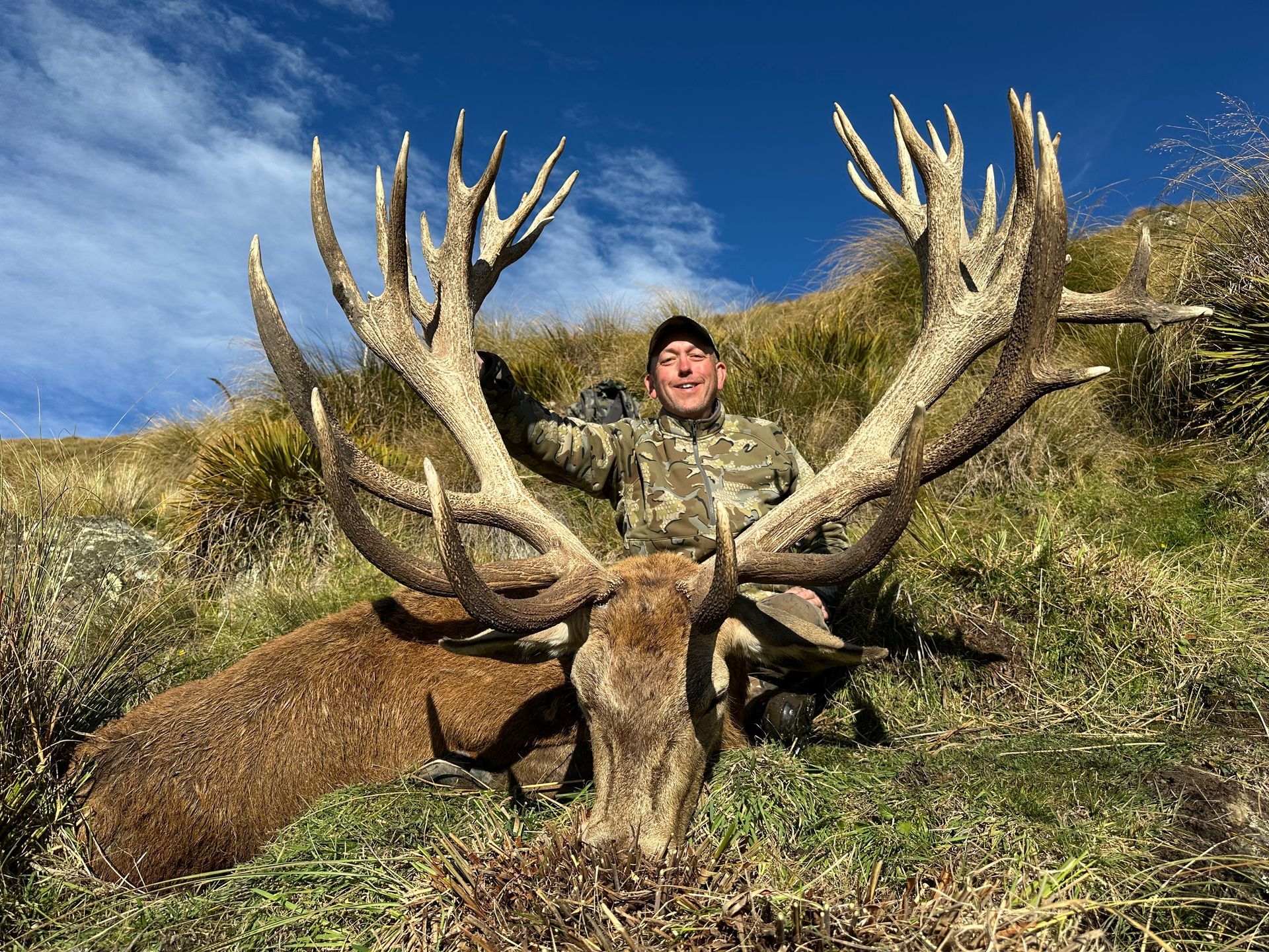 Man in camouflage kneels next to a large deer with impressive antlers, posing in a grassy hillside under a blue sky.