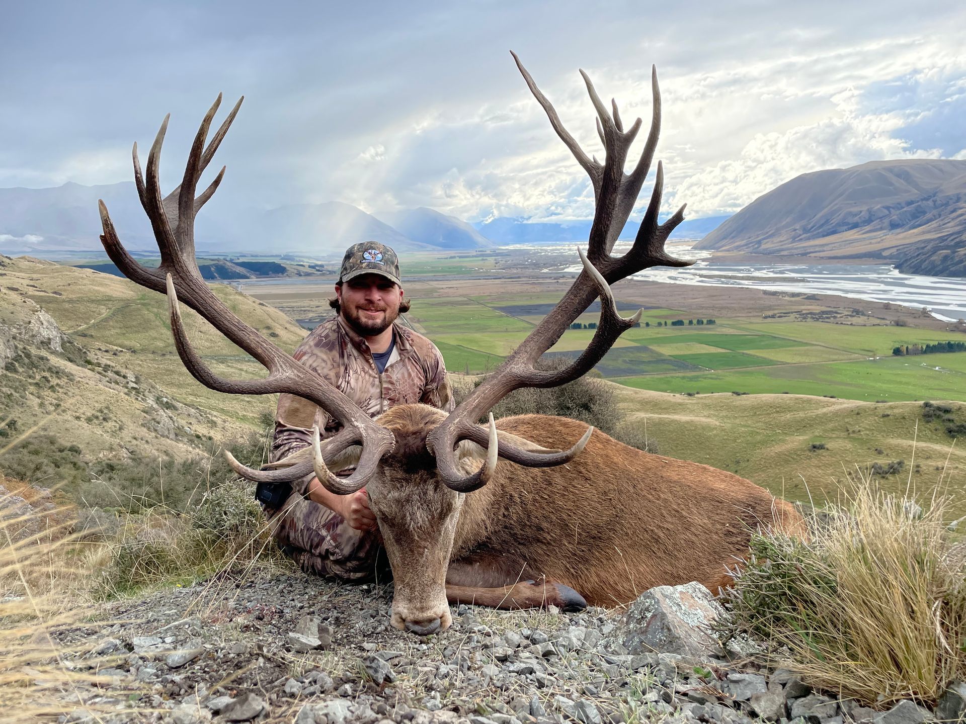 Man in camouflage kneels beside a large red deer with impressive antlers, posing on a hillside with a valley backdrop.