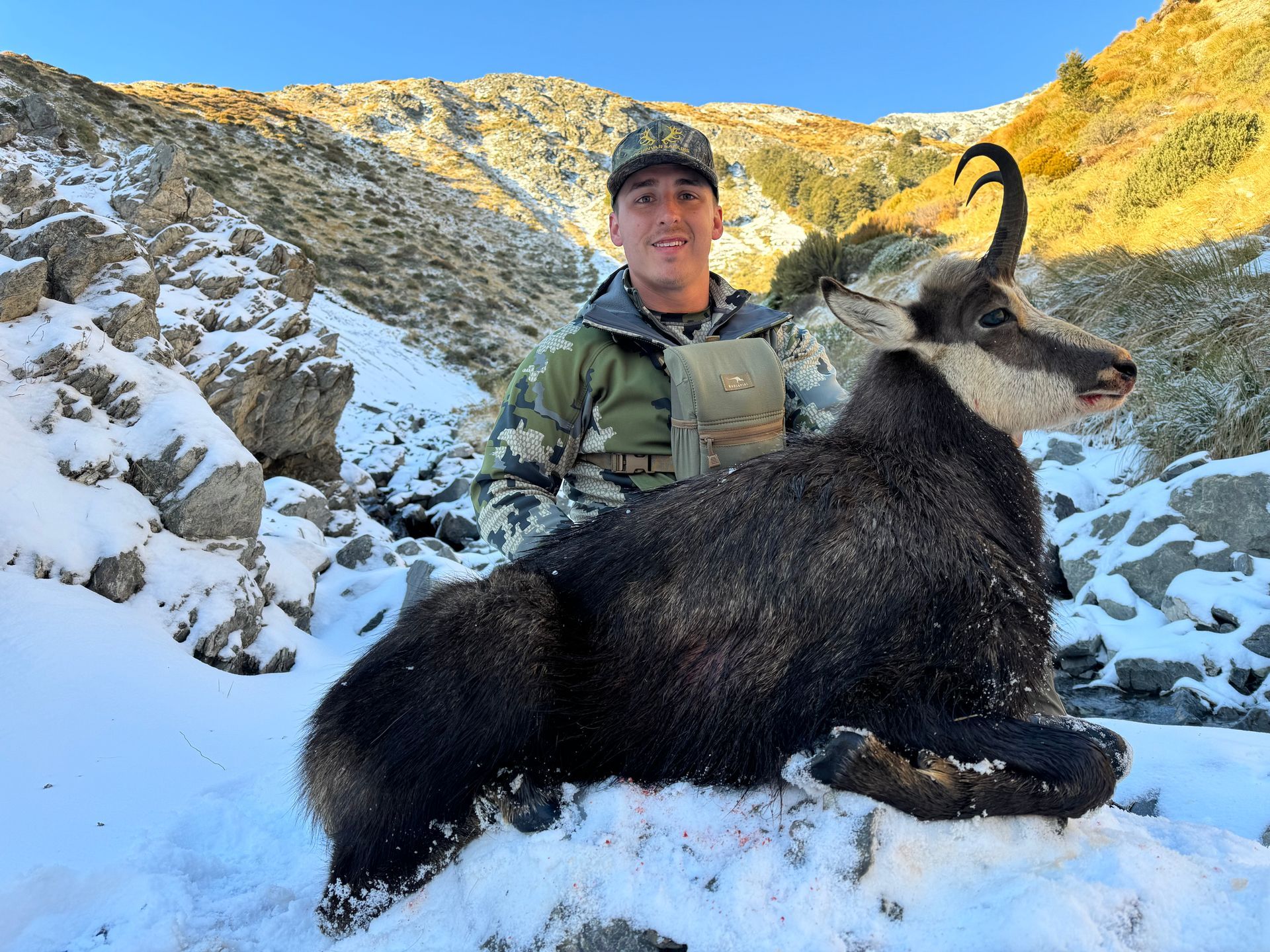 A man in camouflage smiles, posing with a recently hunted black chamois in a snowy mountain setting.