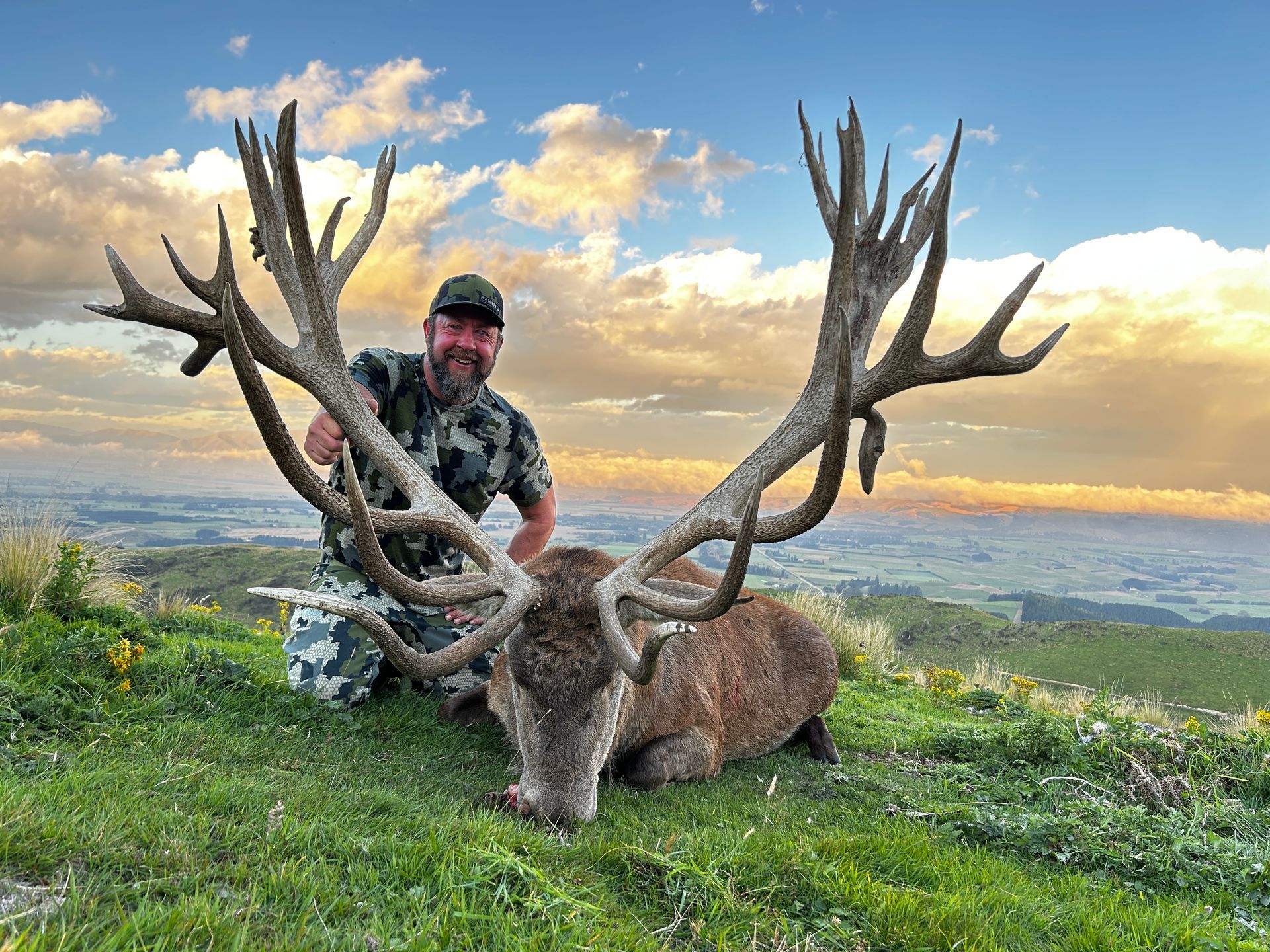 Man in camouflage kneels next to a large stag with enormous antlers, smiling, on a grassy hillside with a scenic view.