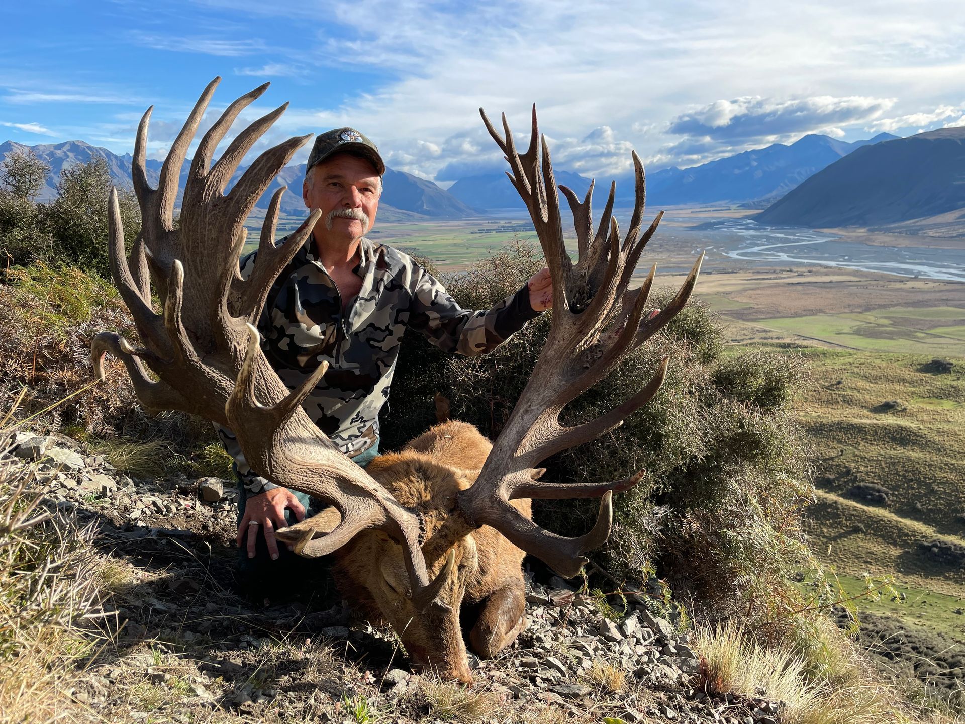 Man in camouflage holds up a massive elk head with large antlers against a backdrop of mountains and a valley.