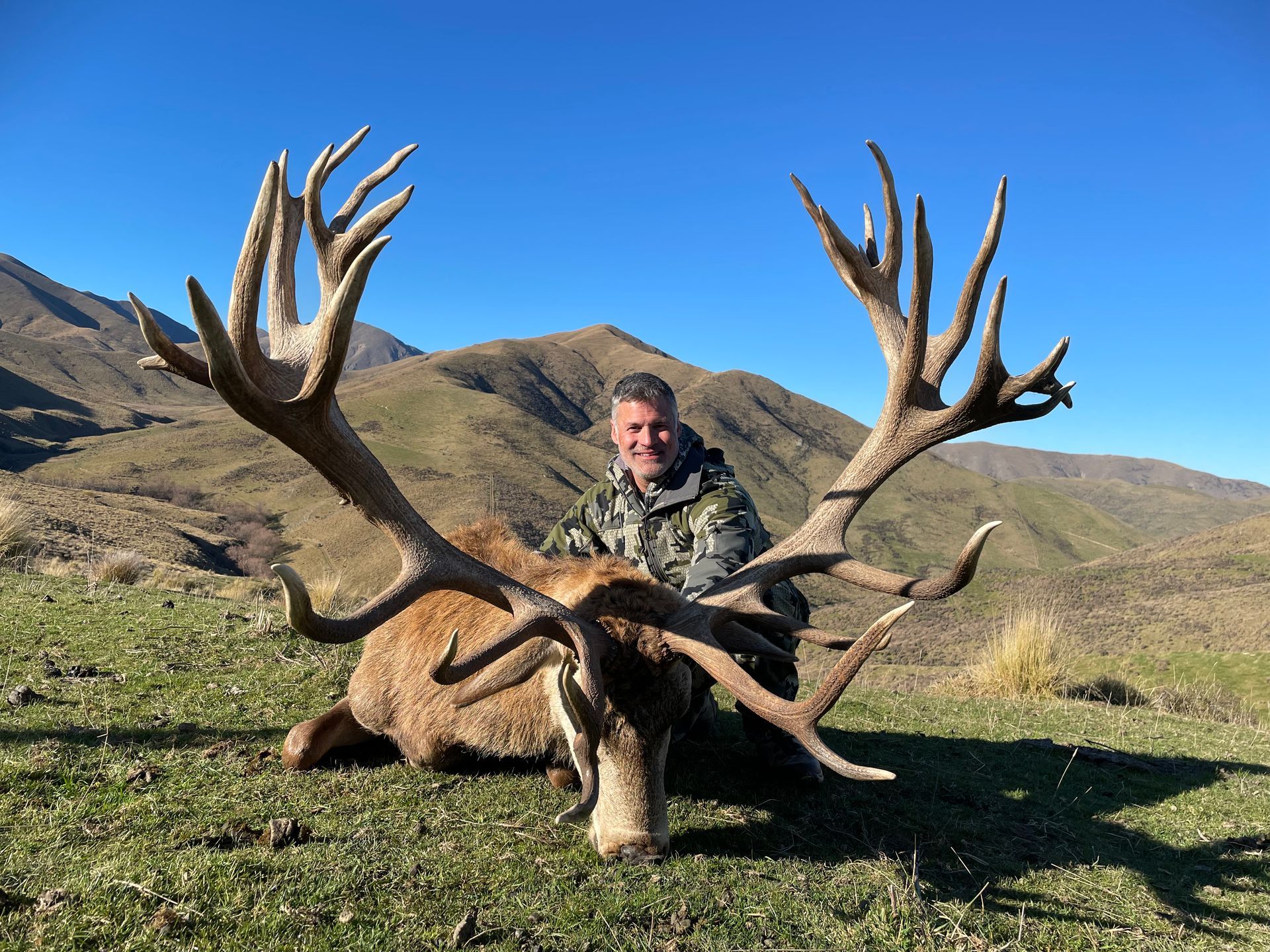 Hunter poses with a large red deer stag in a mountainous, grassy landscape under a clear blue sky. The stag has massive antlers.
