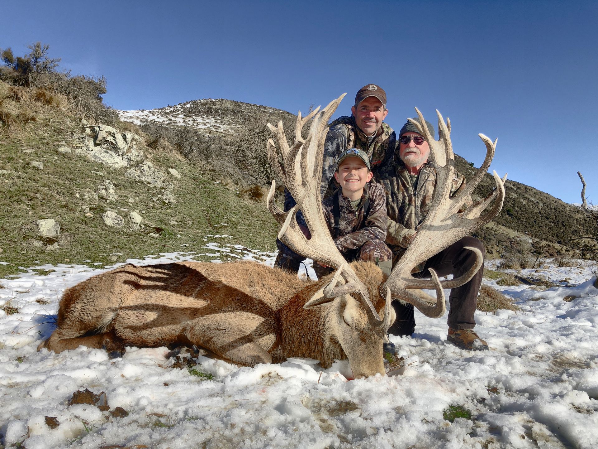 Four people pose with a large dead deer with large antlers in a snowy mountain setting. They are all smiling and wearing camouflage.