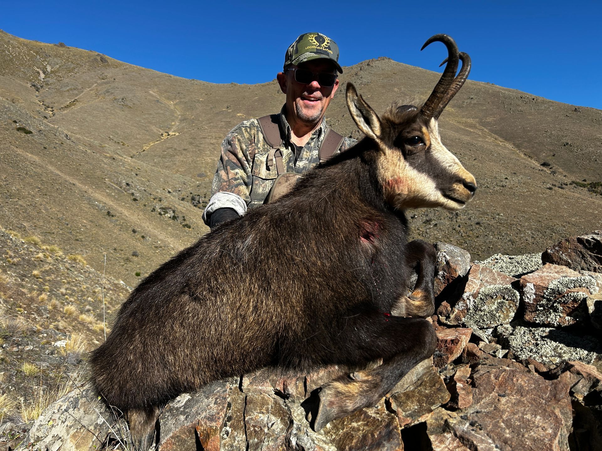 Hunter in camouflage smiles, posing with a dead chamois on a rocky hillside.
