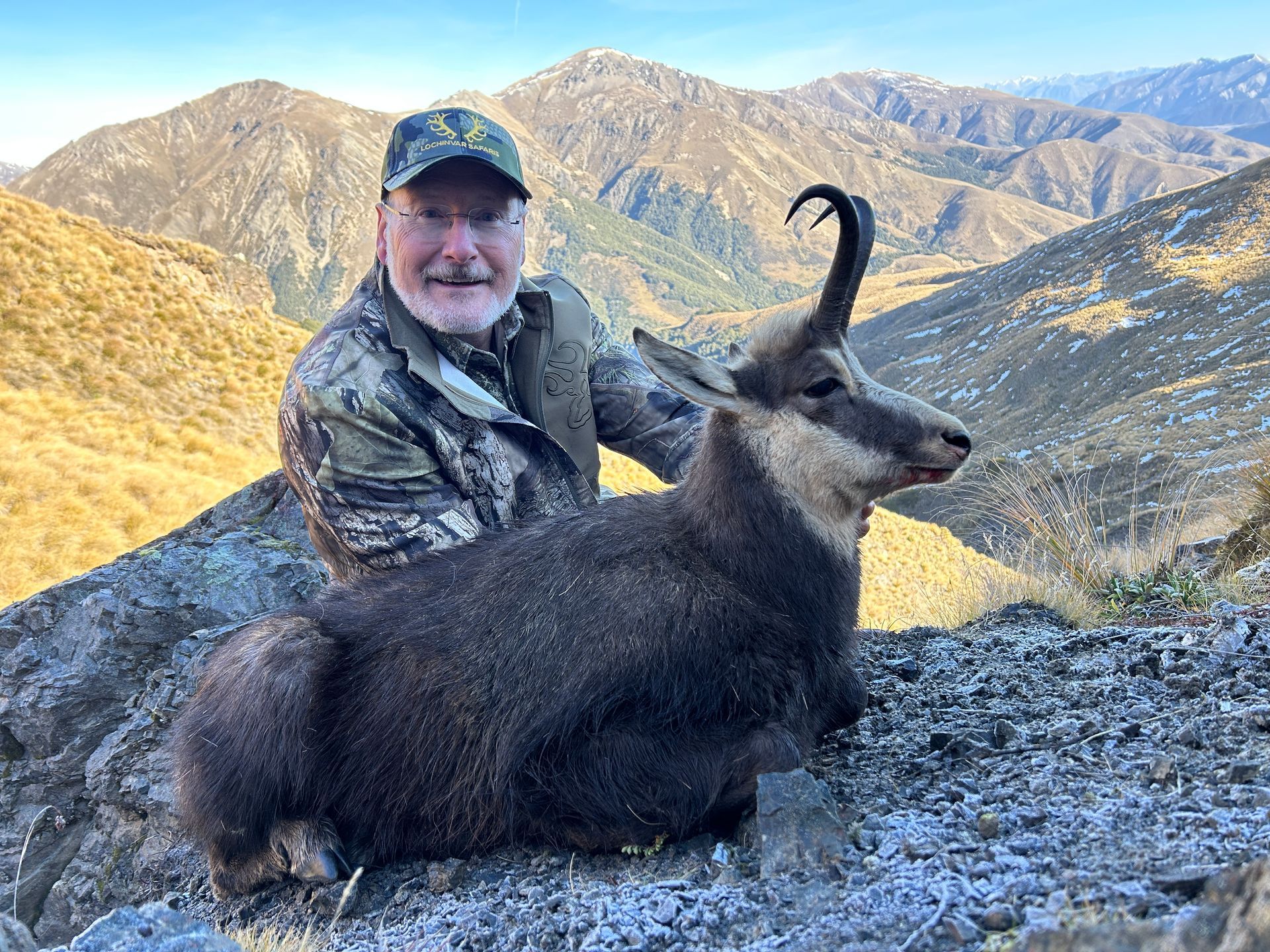 A man in camouflage holds a dark-furred chamois with large curved horns on a mountain with a snowy peak in the background.
