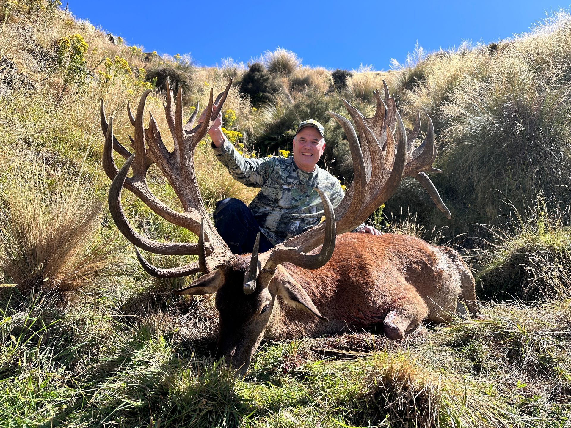 A person sits atop a large red deer carcass with impressive antlers in a grassy, mountainous setting. They are smiling.