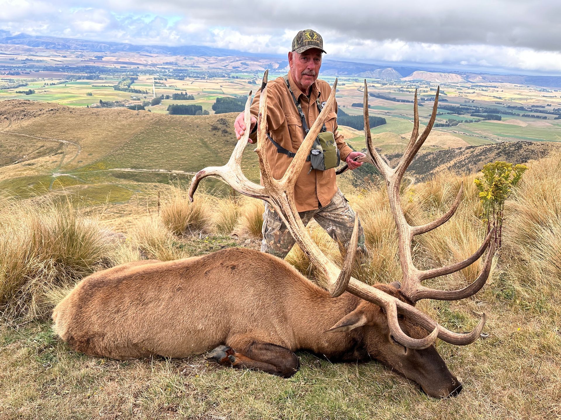Hunter in camouflage holding elk antlers, standing over a harvested elk on a hillside with a distant valley view.