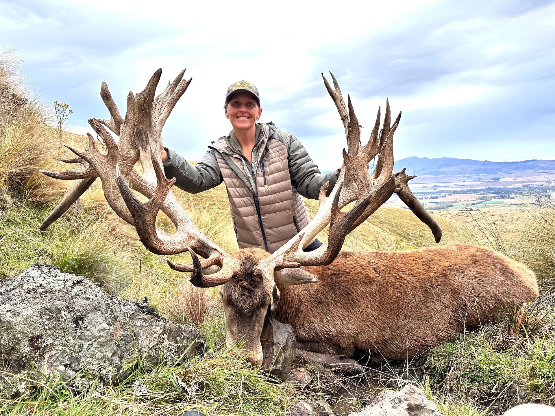 Woman posing with a large red deer trophy in a grassy, mountainous area. She smiles, wearing a jacket and hat.