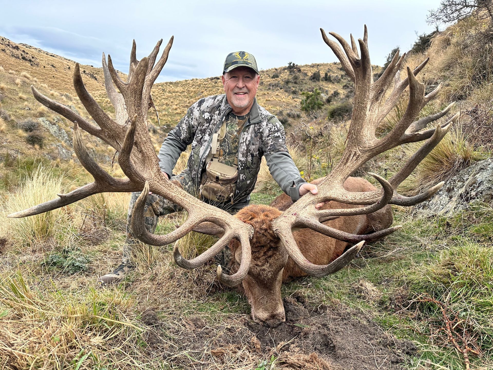 Man in camouflage kneeling beside a large dead deer with enormous antlers in a hillside setting.