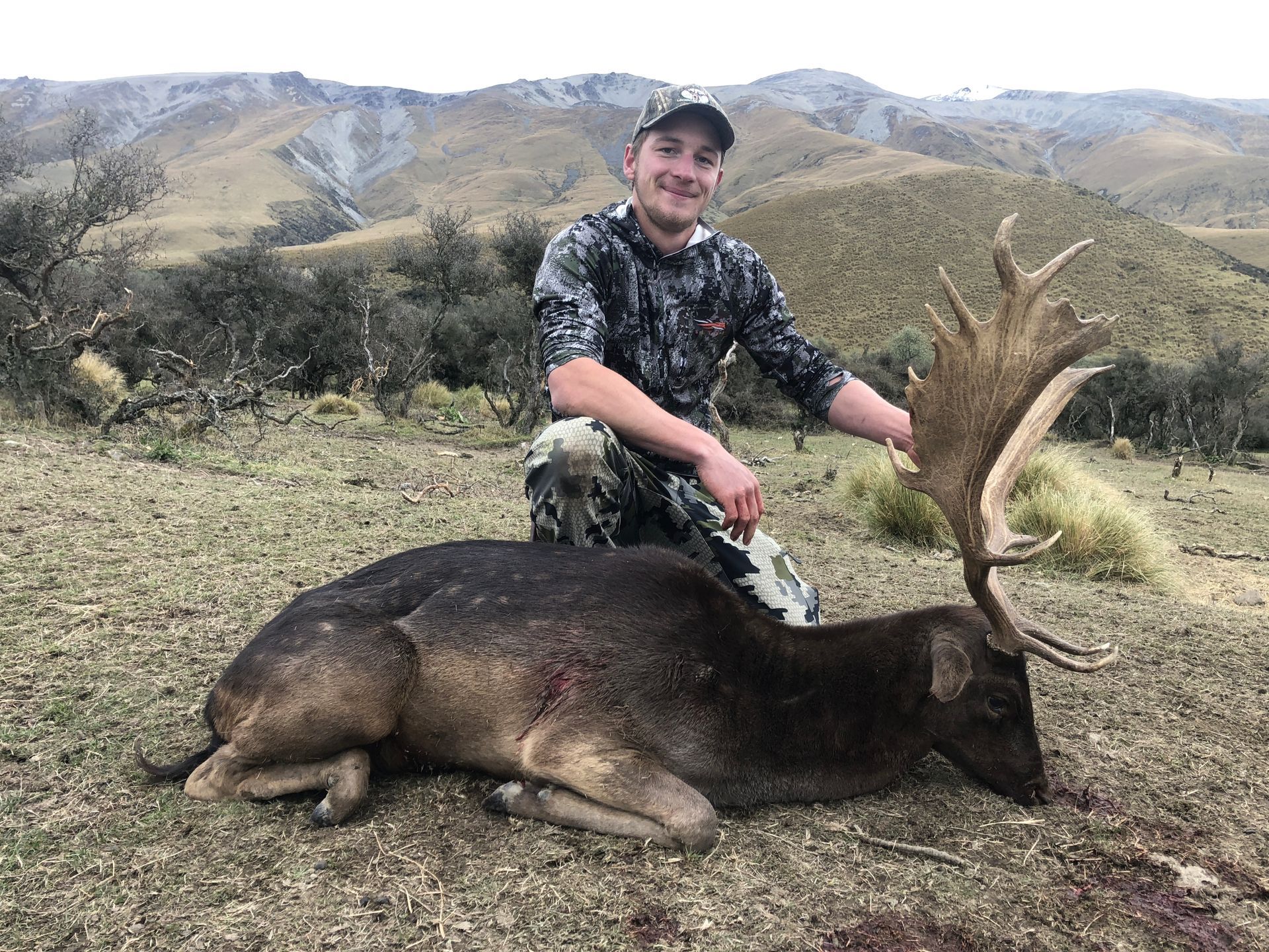Man kneels beside a fallen deer in a mountainous outdoor setting. The man smiles, wearing camouflage. The deer has large antlers.