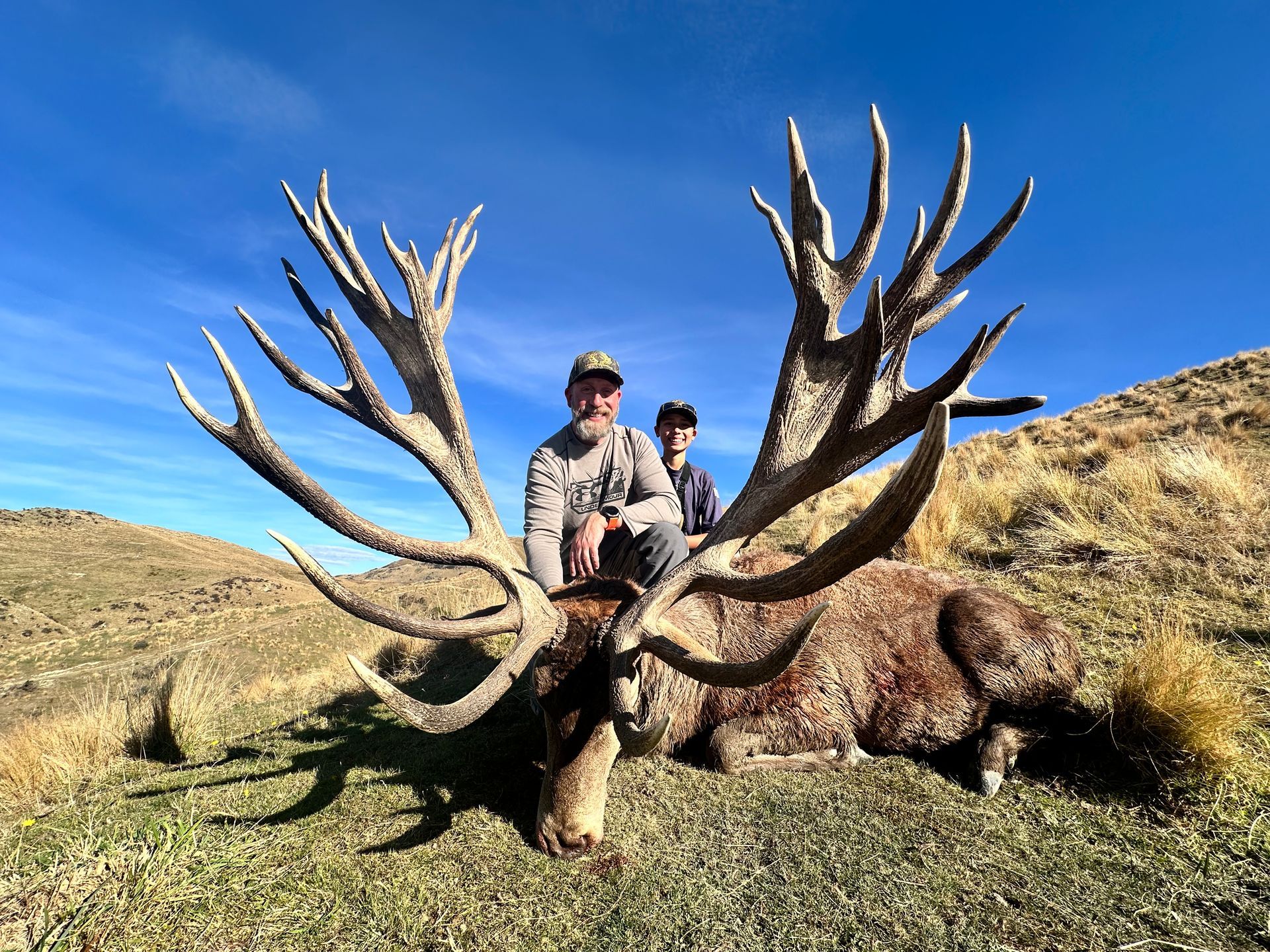 Two people crouch behind a large deer with massive antlers on a grassy hillside. The sky is blue.