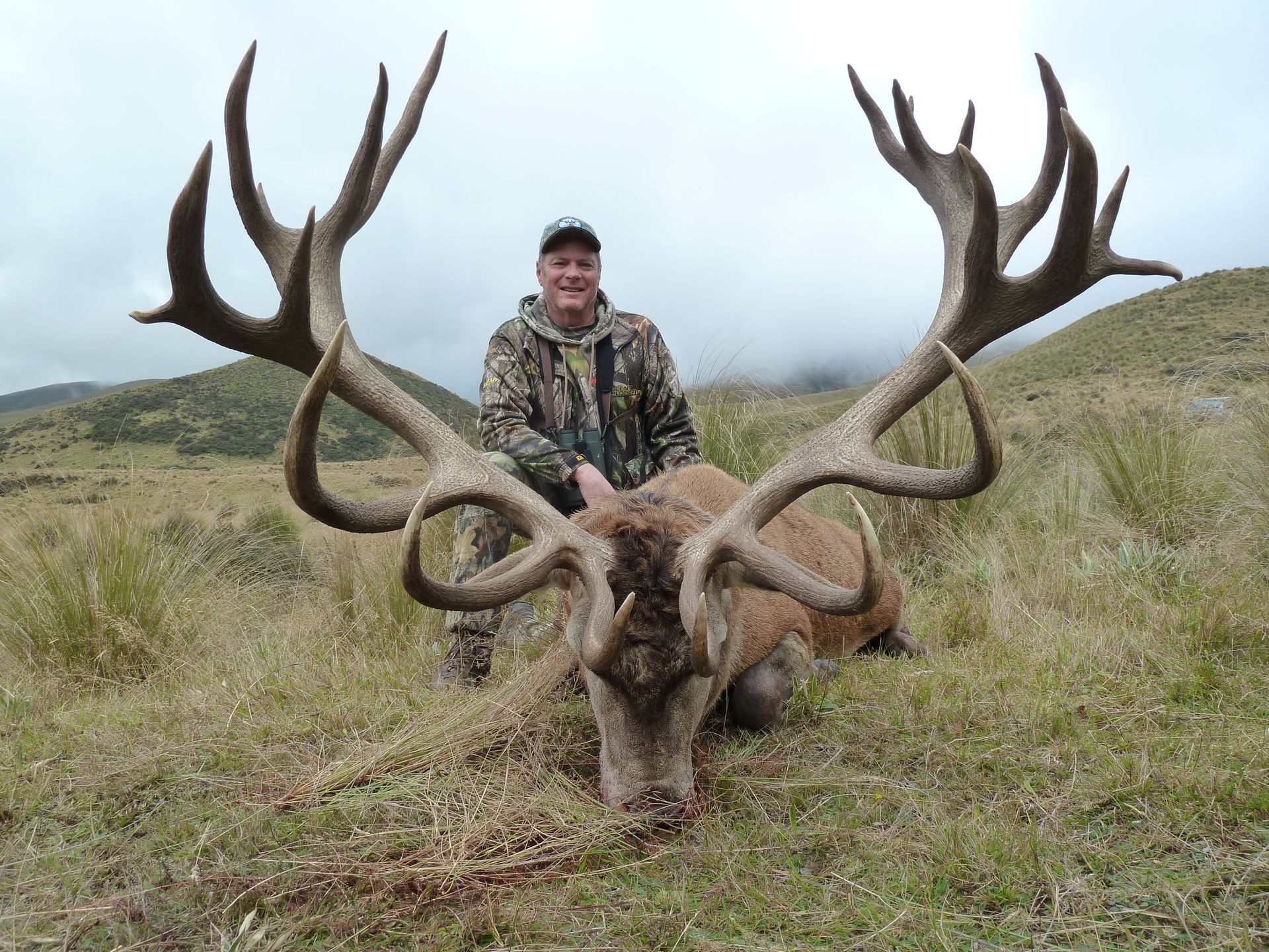 Man in camouflage stands with a large red stag, its massive antlers spread wide, on a grassy hillside.