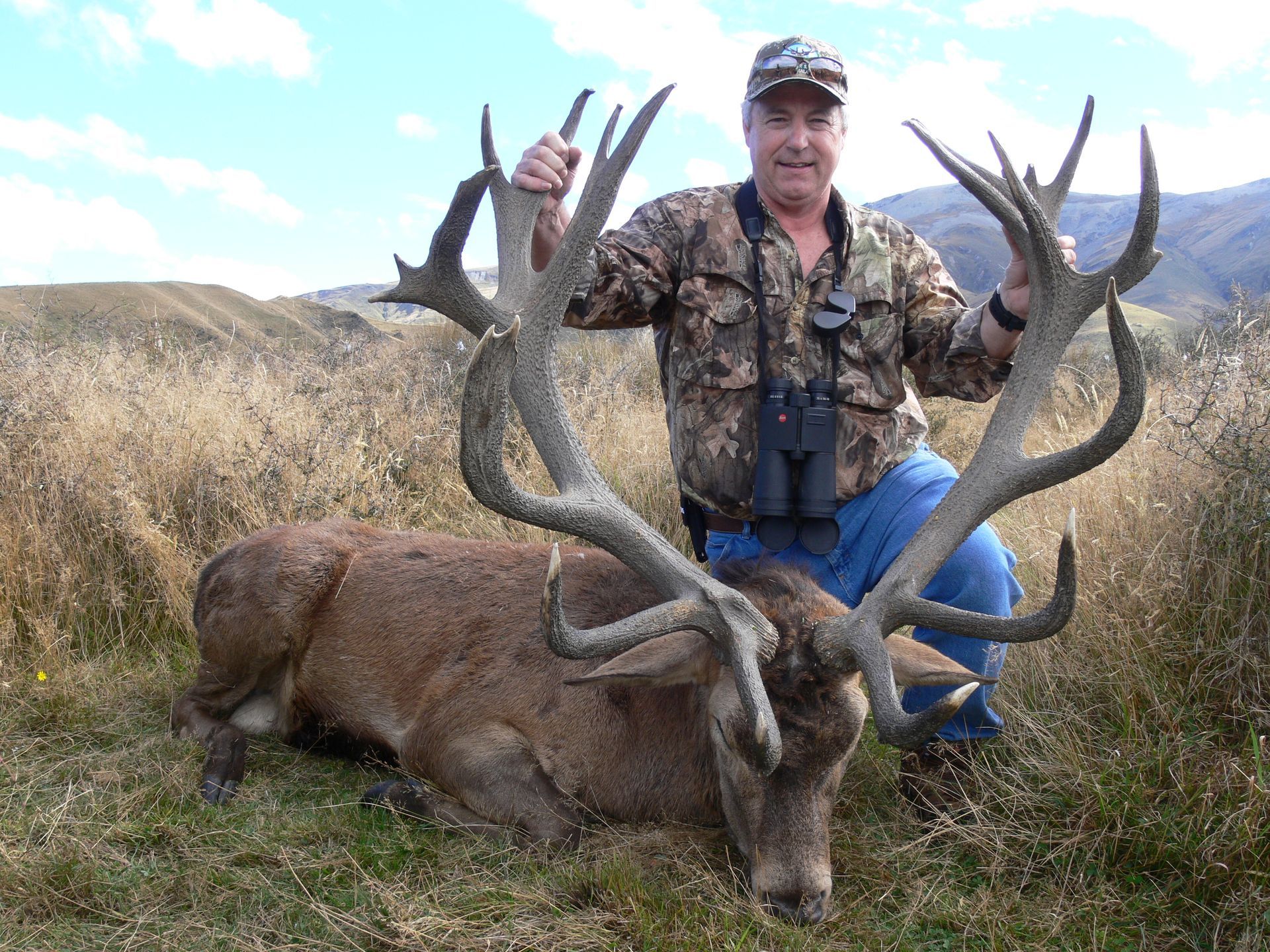 Man kneels beside a large red deer stag with impressive antlers in a grassy field. He wears camo and smiles.