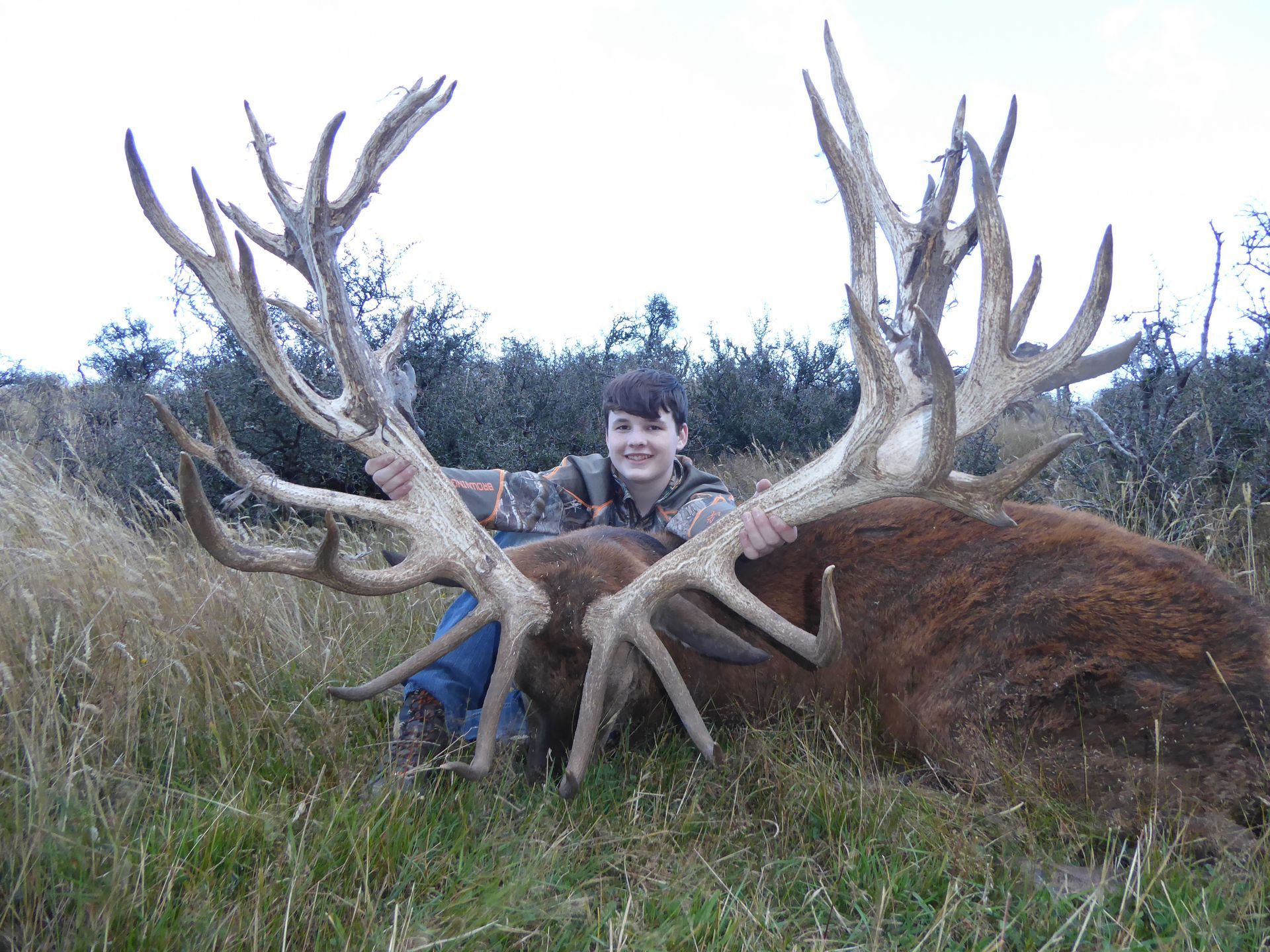 Young person in camouflage holding large antlers of a dead red deer in a field; they are smiling.