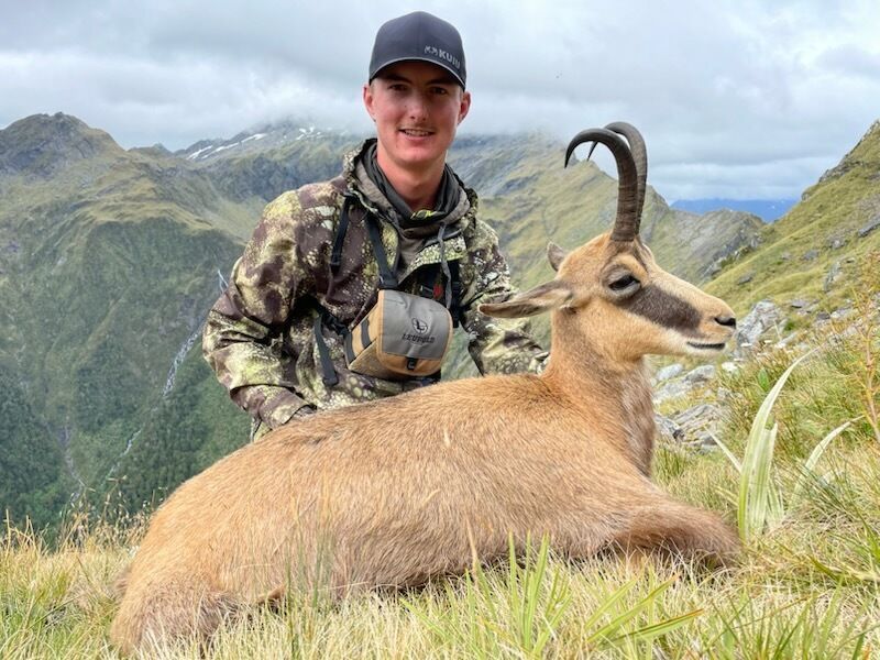 Hunter in camouflage gear smiles beside a chamois he has harvested in a mountainous landscape.