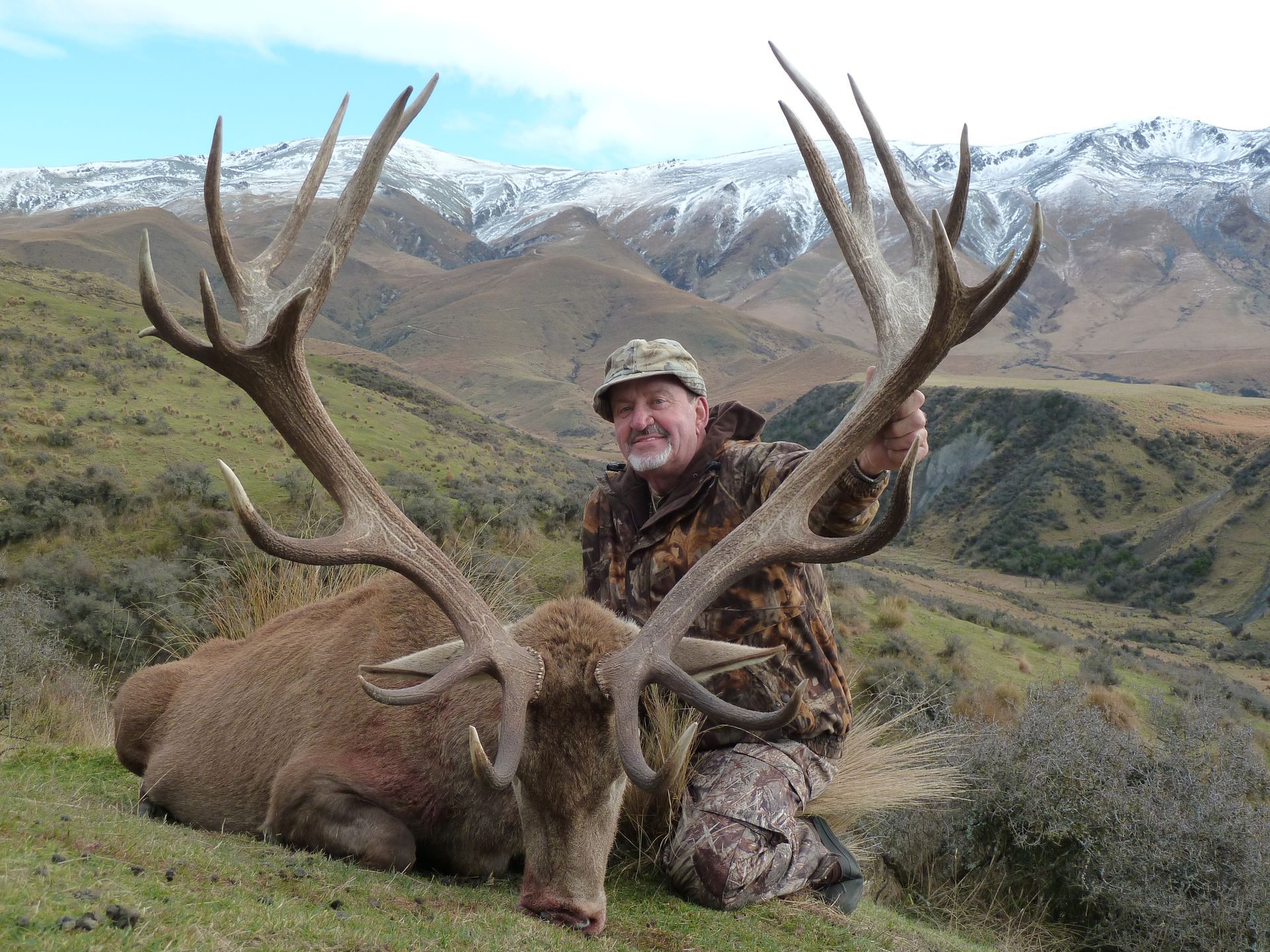 Man in camouflage kneeling beside a large dead deer with impressive antlers in a mountainous landscape.