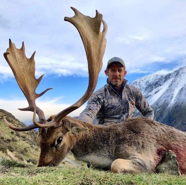 A man in camouflage poses next to a large, dead deer with impressive antlers in a mountain setting.