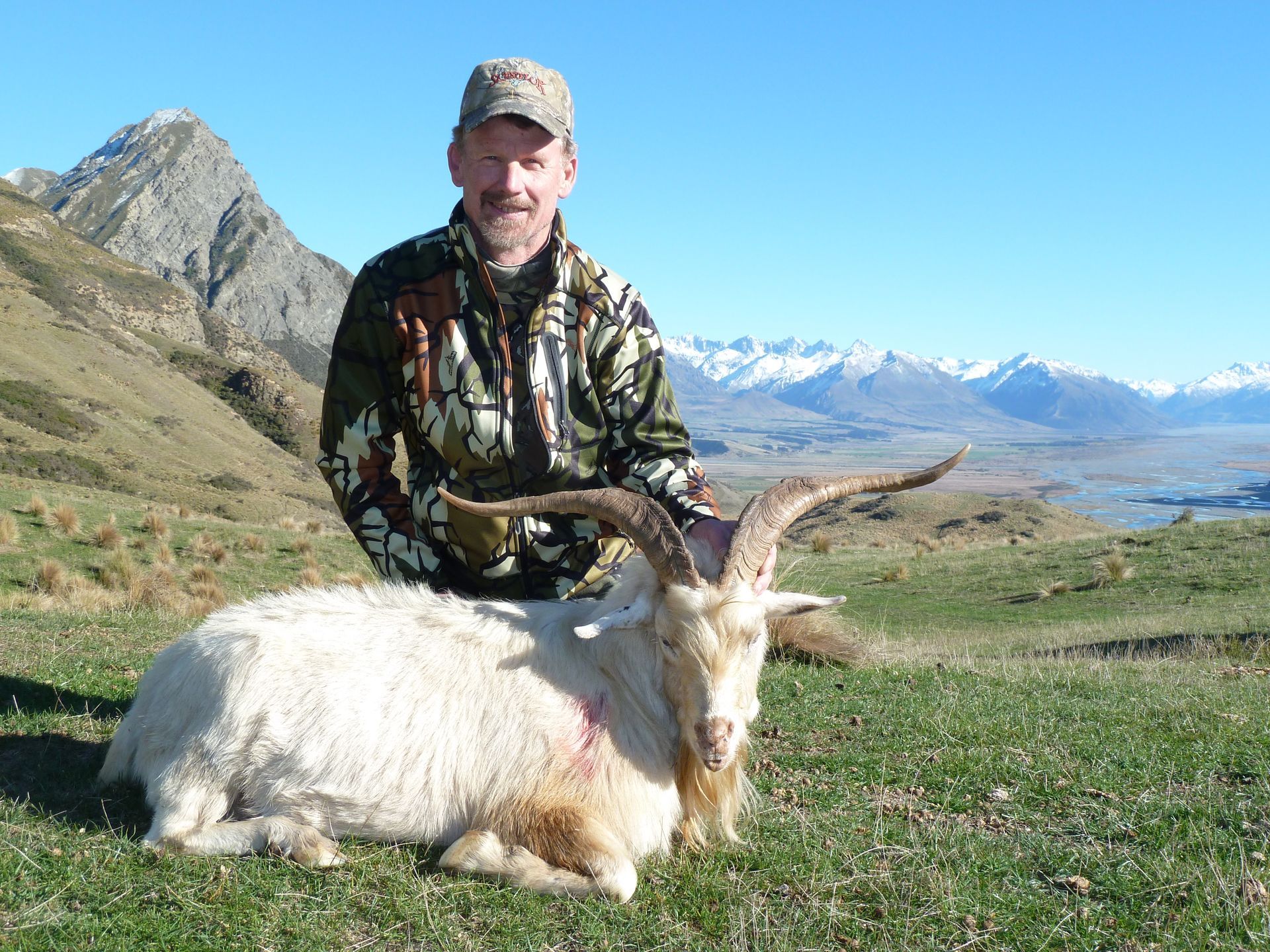 Man in camouflage with a large white goat he has hunted, mountains in the background.