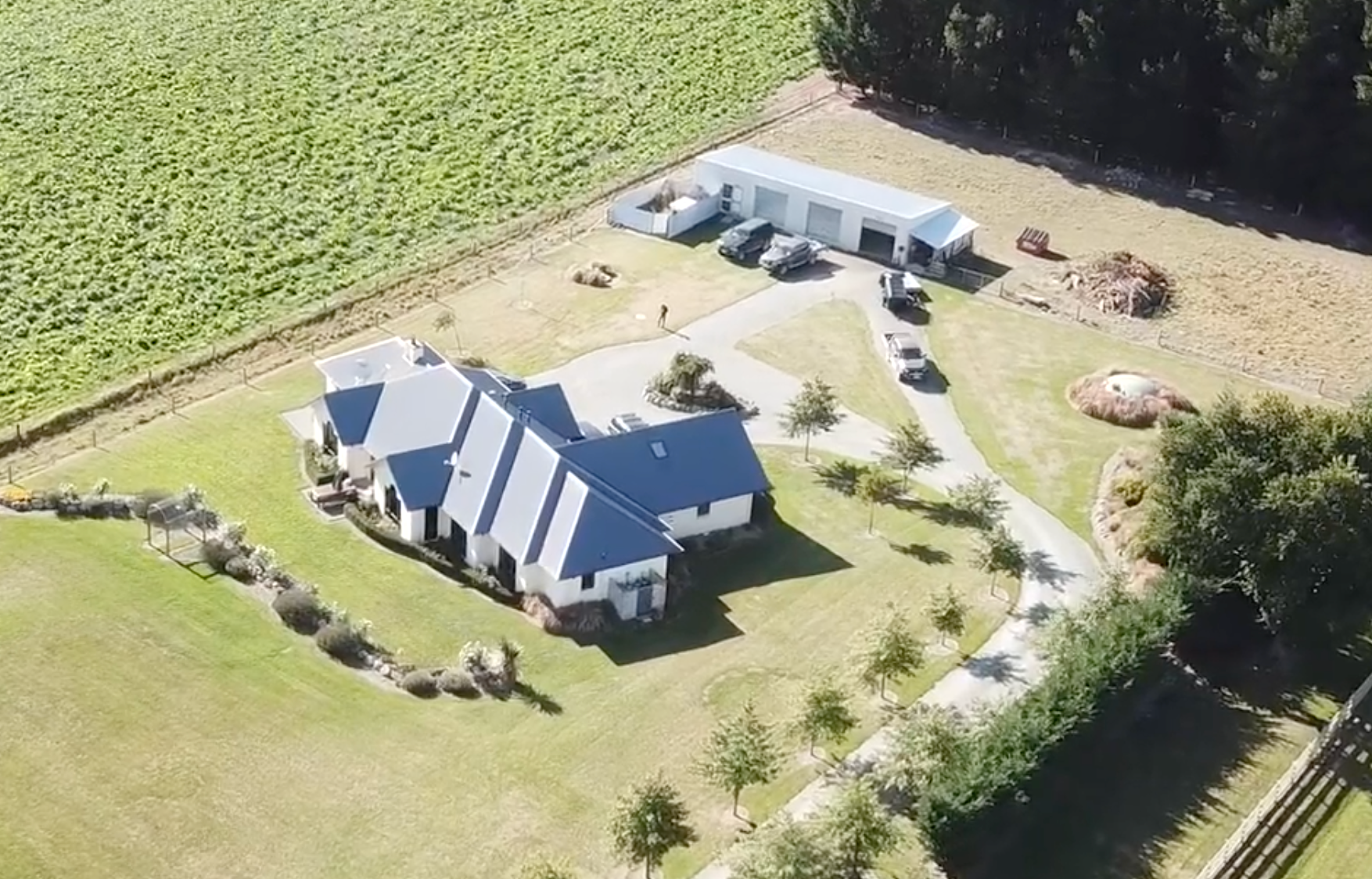 Aerial view of a house with a dark roof, a driveway, and a shed. Green fields surround the property.