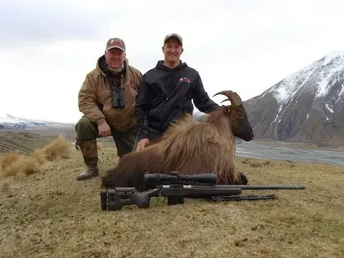 Two men pose with a dead Himalayan tahr in a mountain landscape. A rifle rests in front of the animal.
