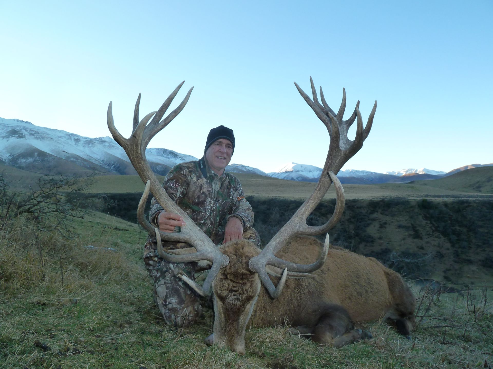 Man in camouflage kneeling beside a large harvested red deer with impressive antlers, mountains in background.