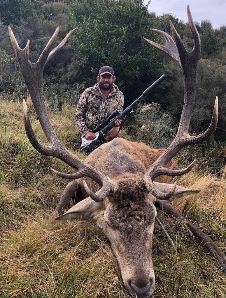 A man in camouflage poses with a large dead stag in a grassy area. He holds a rifle, smiling.