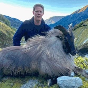 Man in blue jacket poses with a large, shaggy Himalayan tahr he appears to have hunted, mountains in the background.