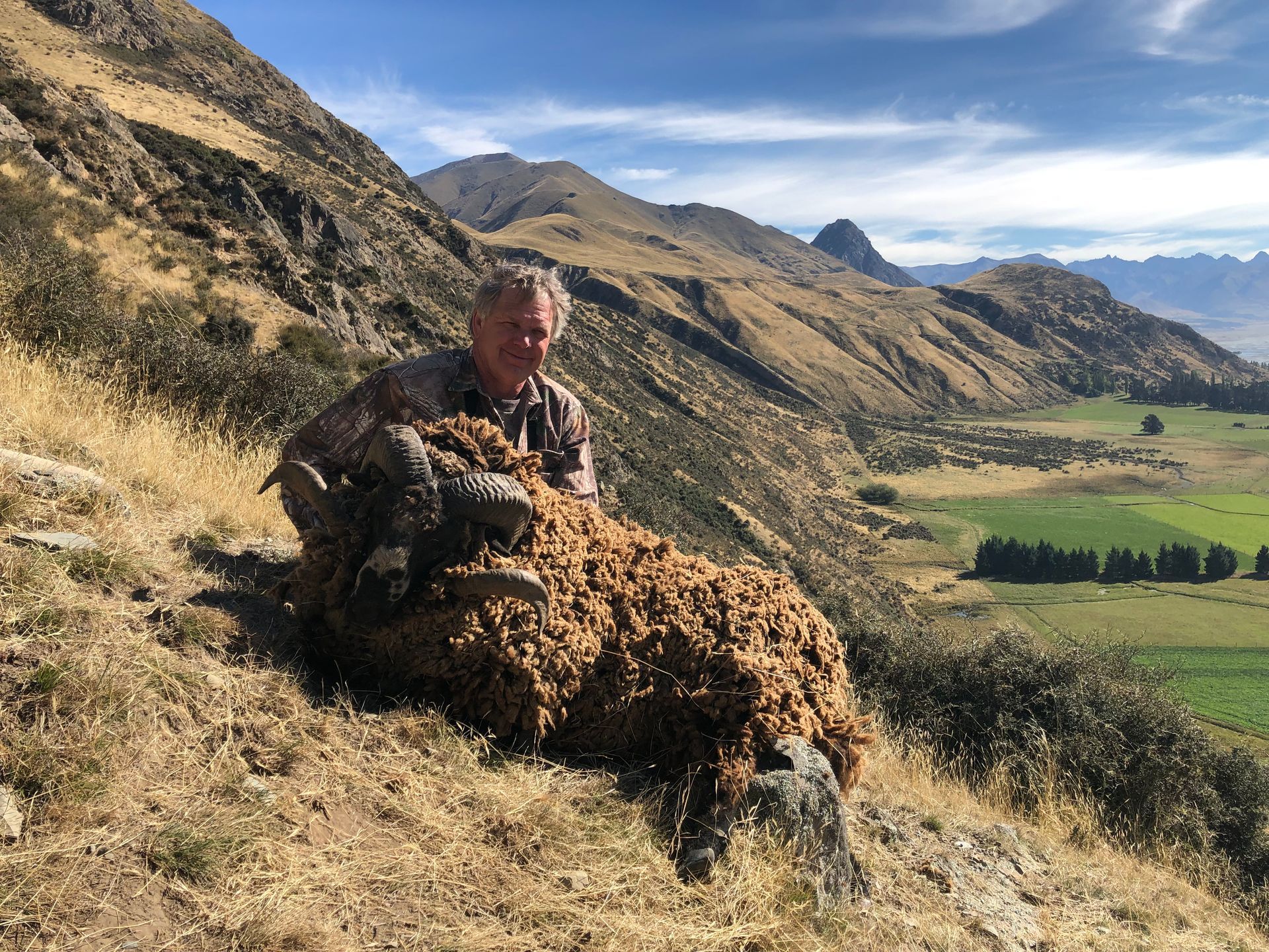 Man kneeling with a large, brown-haired animal in a mountainous, grassy setting. The man smiles at the camera.
