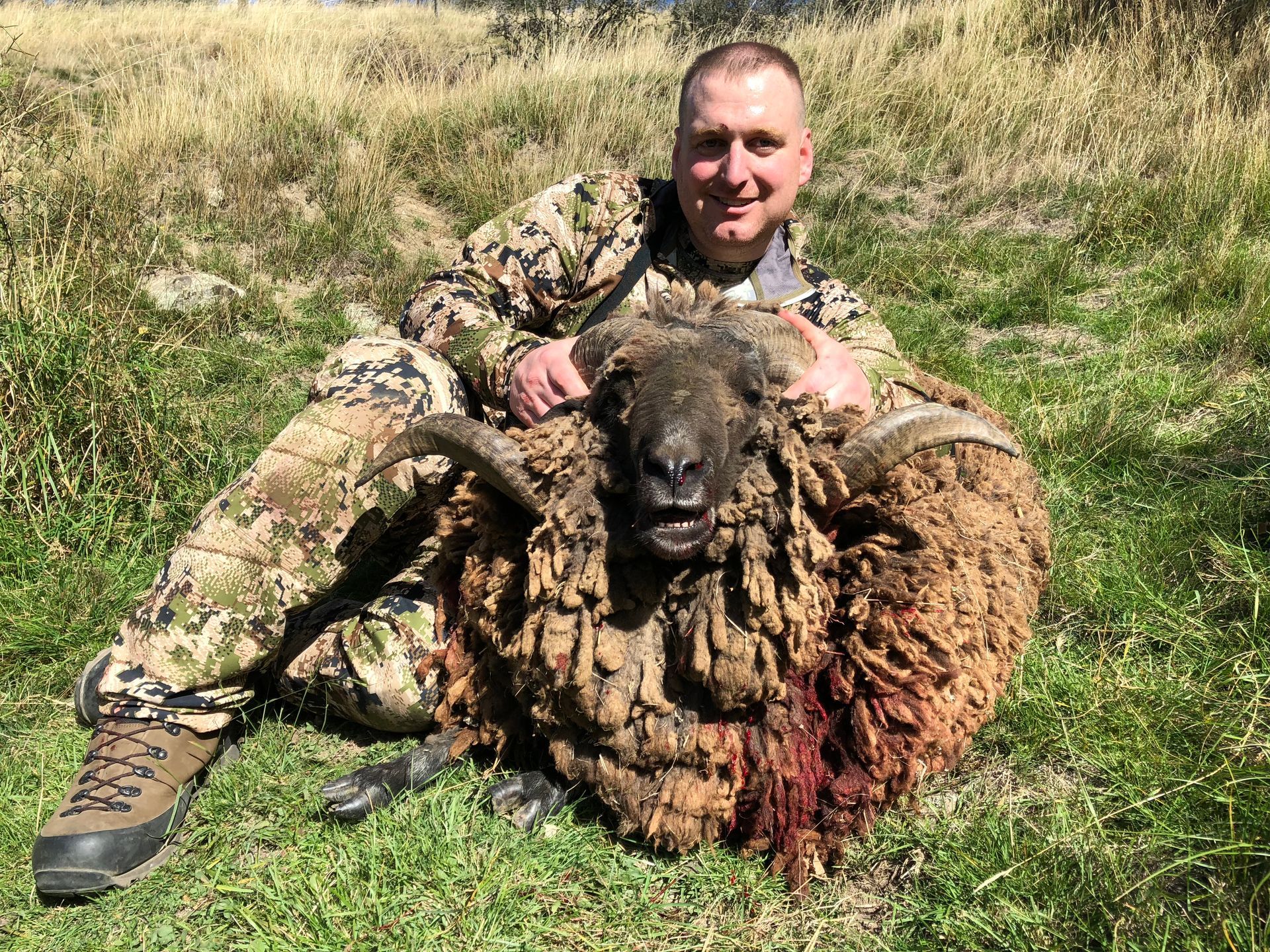 Man in camouflage, smiling, kneels beside a large sheep with blood on its wool in a grassy outdoor setting.