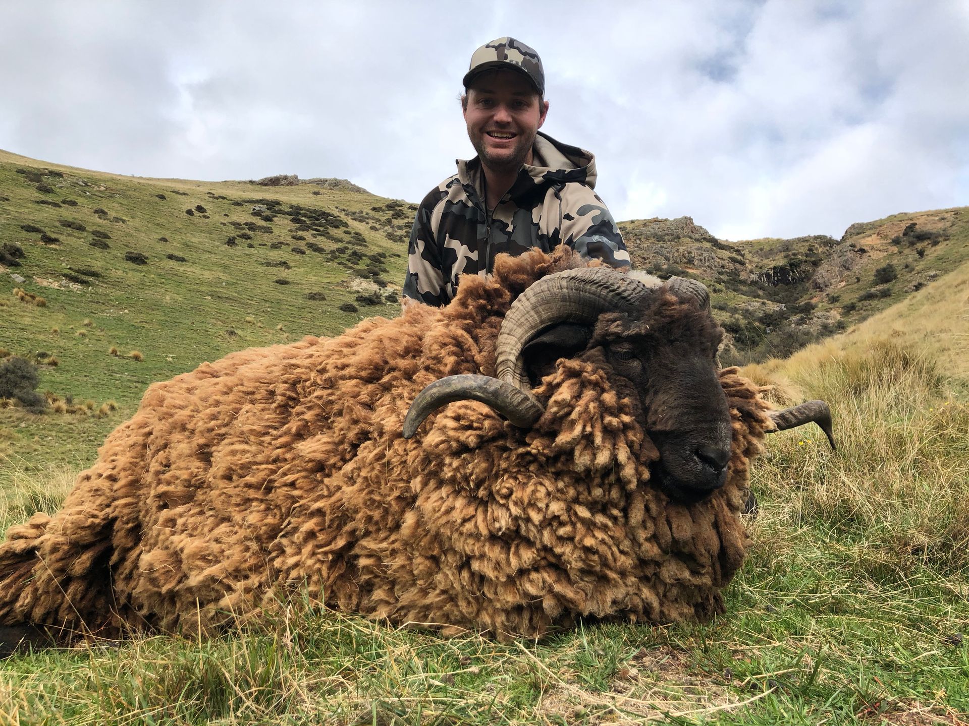Man in camouflage posing with a large, brown-wooled ram in a grassy, mountainous setting. The man is smiling.