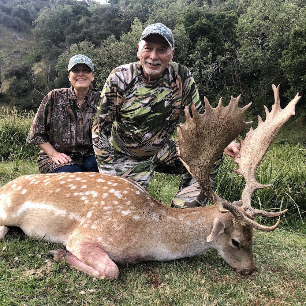 A man and woman pose with a large, spotted buck. They are outdoors in a grassy area. The man is wearing camouflage clothing and smiling.