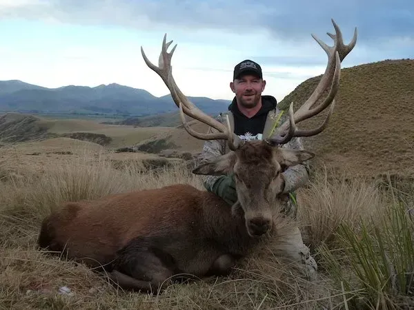 Man smiling next to a large, antlered deer he has hunted, set against a mountain landscape.