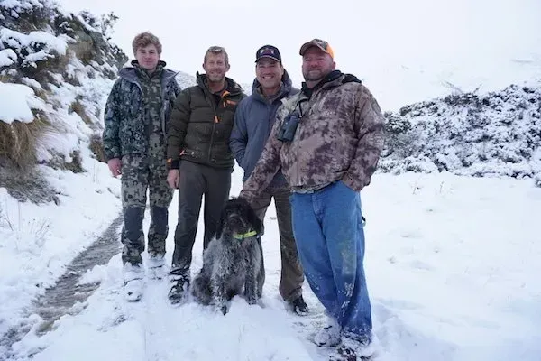 Four men and a dog in snowy landscape. The dog sits in front of the group, with one man petting it.