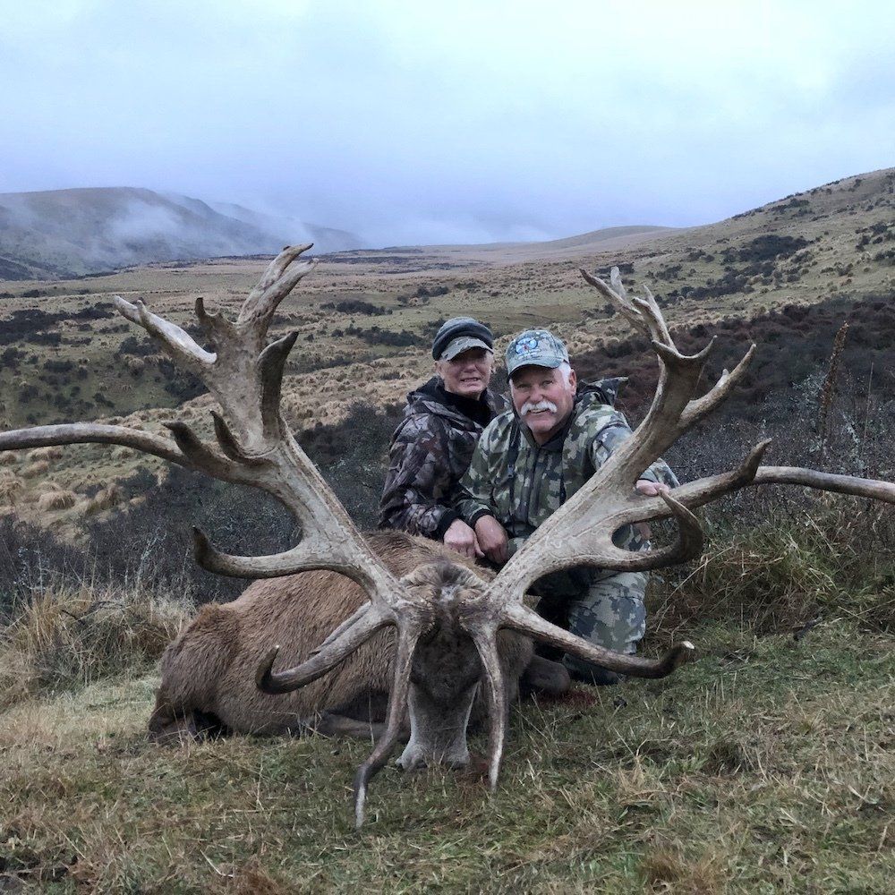 Two people pose next to a large dead deer with impressive antlers in a grassy, mountainous area. They're smiling, and the sky is overcast.