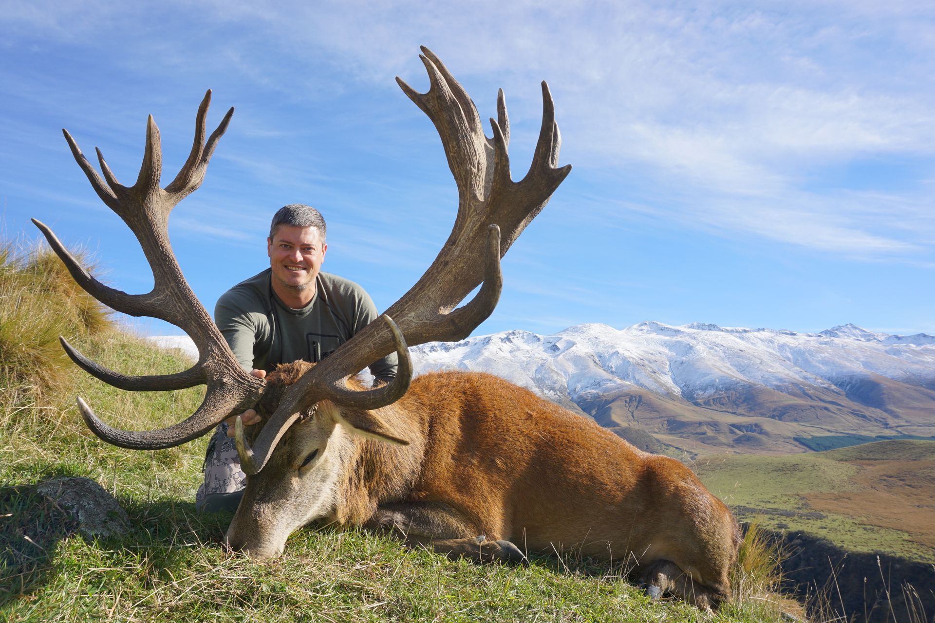 Man kneeling beside a large, dead red deer with impressive antlers, posing in a grassy field with mountains in the background.