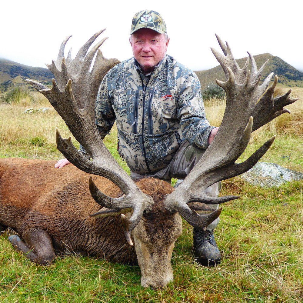 Man in camouflage poses next to a large dead red deer with impressive antlers in a grassy field.