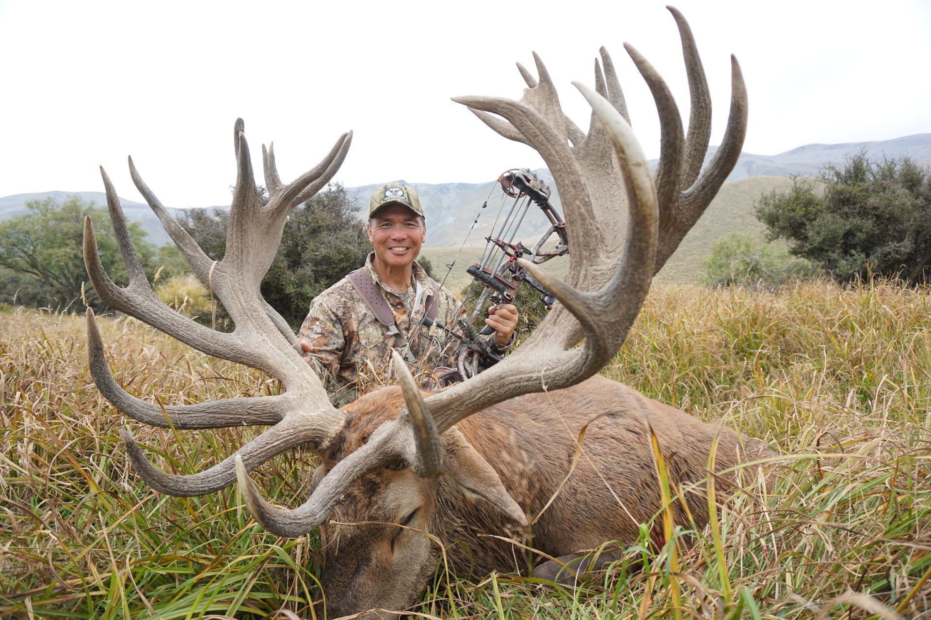 A hunter in camouflage gear smiles, holding a bow over a large dead elk with huge antlers in a grassy field.