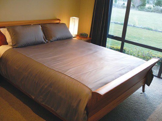A well-made bed with gray bedding in a bedroom next to a large window overlooking a grassy area.