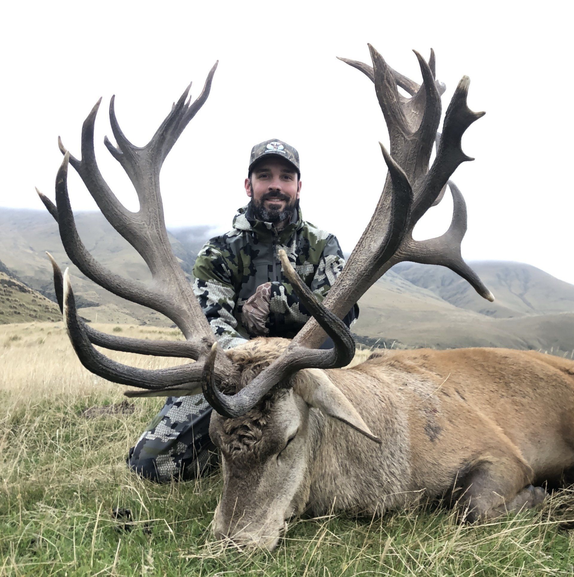 A man kneels beside a large dead red deer in a grassy field, mountains in the background. He wears camouflage and holds a small object with a pleased expression.