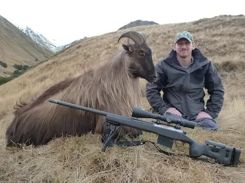 Hunter kneels beside a dead Himalayan tahr on a hillside, with a rifle resting in front of them. Snowy mountains in the background.