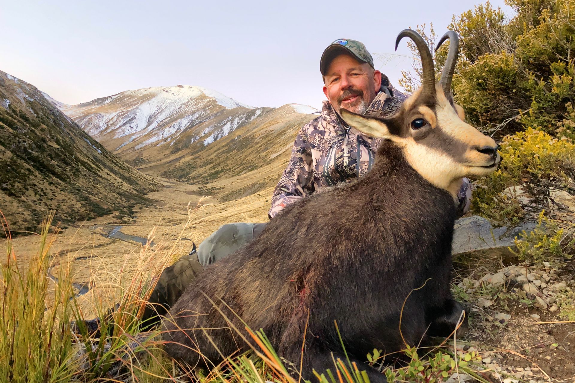 Man in camo with a freshly hunted chamois in a mountain landscape; the chamois is black and has large horns.